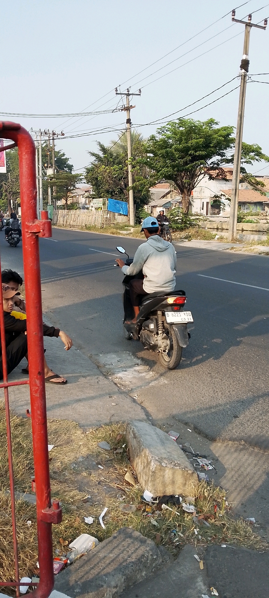 The image captures a scene on a street in Bekasi, Indonesia. In the foreground, a red metal structure is partially visible on the left, with dry grass and scattered litter around its base. Two individuals are seated on a concrete curb to the left, one wearing a yellow shirt and black pants, the other a tan cap and black shirt. A lit cigarette is held by the person on the right. A person wearing a grey hoodie and a light blue cap is riding a black motorcycle away from the camera, their body turned to the left. Another person on a motorcycle is visible further down the street, also moving away. The road is paved, and a drainage ditch with more litter runs alongside the curb. Power lines are strung across poles in the background, above a row of buildings and trees. The sky is clear and bright.