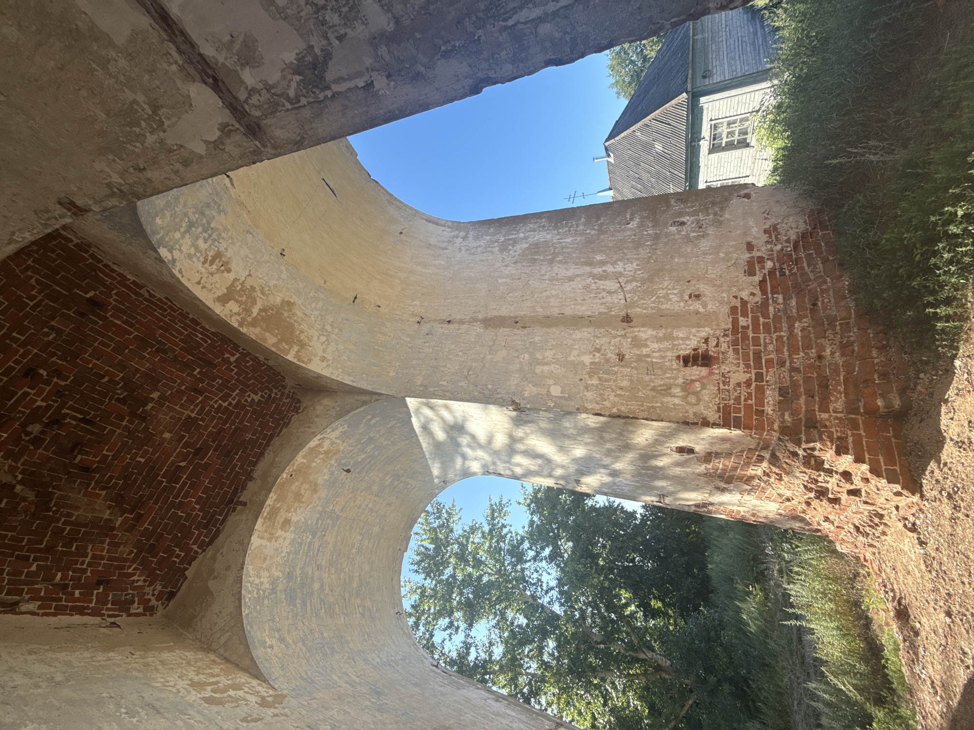 An upward angle shot captures a view through two large, weathered stone arches against a clear blue sky. The interior of the arches reveals exposed red brickwork on the ceiling, suggesting an old, possibly abandoned structure. The rough texture and peeling paint on the stone pillars indicate significant age and decay. To the left, the arch frames a lush green tree. To the right, beyond the second arch, a wooden building with a grey, corrugated roof and a single window is visible, partially obscured by tall green grass. The ground in the foreground appears to be a mix of sand and dirt, with some sparse vegetation. The scene has a somber, derelict quality, hinting at a forgotten past.