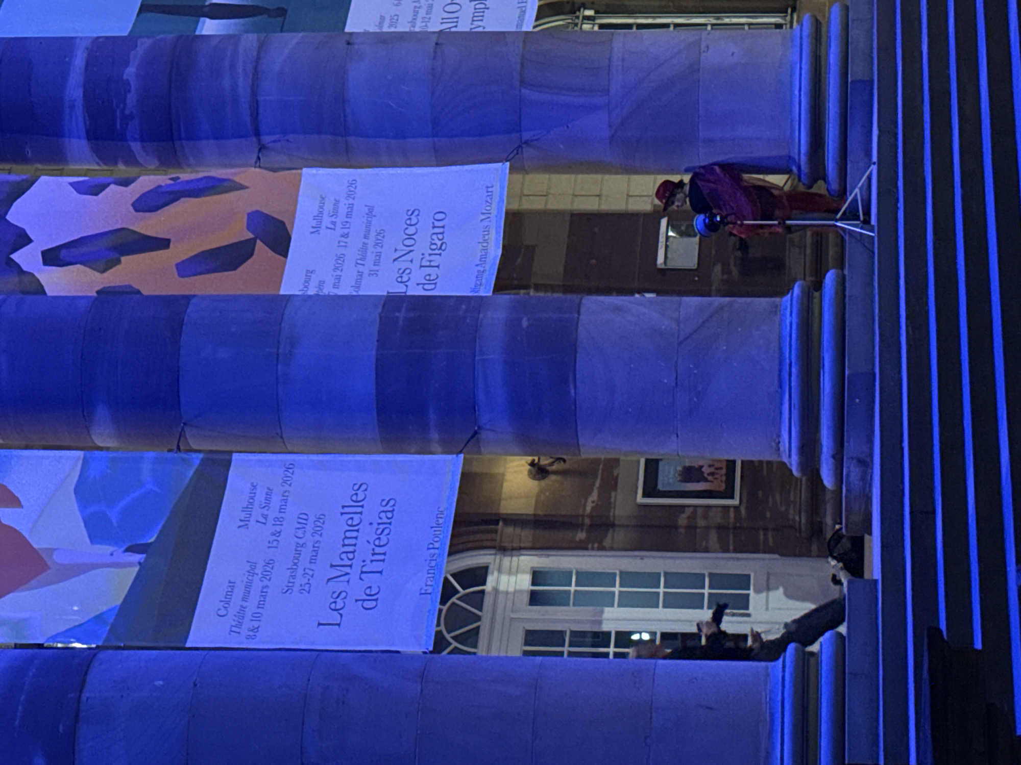 The image depicts an exterior building facade, likely in Strasbourg, France, due to the text on the displayed banners. The facade consists of a series of large, cylindrical columns that are illuminated with a blue light. Banners are hanging between the columns, advertising performances of operas, including "Les Mamelles de Tirésias" by Francis Poulenc and "Les Noces de Figaro" by Wolfgang Amadeus Mozart. The banners list performance dates in Colmar, Mulhouse, and Strasbourg in March and May of 2026. Two individuals can be seen on the right side of the frame, one appearing to be standing on a ladder near the middle columns and the other is positioned near the bottom columns.