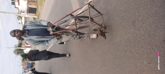A man in a light-colored jacket and light-colored pants stands on a road next to a red, metal frame that appears to be a modified bicycle. The bicycle has a wheel within the frame. Another person in a dark jacket walks toward the camera. Buildings and a utility pole are visible in the background. The road surface is dark. The text "police kombo" is at the bottom right corner.