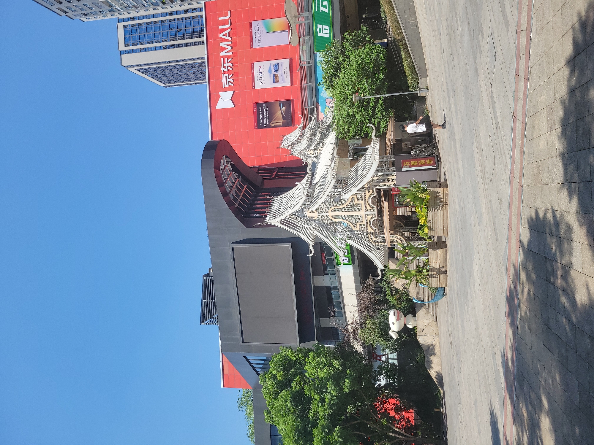 This is a ground-level, eye-level, full shot of a plaza in front of a modern commercial building. The building's facade is predominantly red with "京东 MALL" (JD MALL) written in white Chinese characters and the JD logo. A large, dark, rectangular screen is visible on the building's facade. To the left of the screen is a large black structure with a curved red entrance. To the right of the red facade are taller, modern apartment buildings. In the foreground, a stylized, ornate white pavilion with multiple tiered roofs sits in the plaza. To the left of the pavilion, a white, cartoonish dog statue with a red collar is positioned on a raised, rocky mound. Green trees and lush foliage are scattered throughout the scene. A single individual, wearing a white t-shirt and black shorts, stands in the plaza, facing away from the camera. The plaza floor is paved with grey tiles and has a red and grey striped border. The sky is clear and blue.