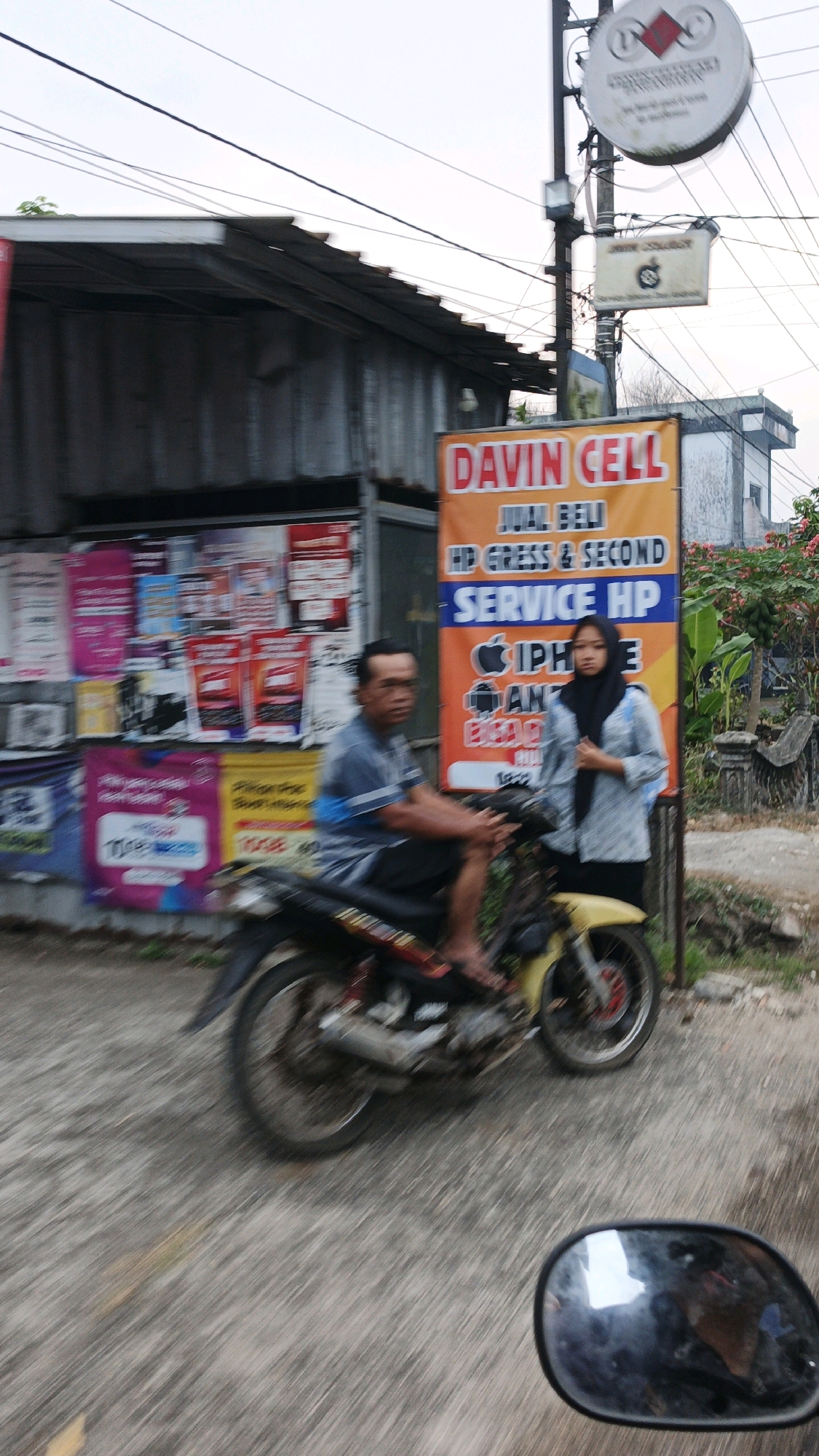 A man is riding a motorcycle on a road in Cibenda, Indonesia. He is wearing a blue and grey striped shirt and black shorts. To his right, a young woman wearing a black hijab and a grey and white patterned shirt stands near a sign for "Davin Cell" which advertises phone sales and services. The sign is orange and white and features the logos of Apple and Samsung. Behind the man, a building with a metal roof and corrugated metal walls is visible, covered in colorful posters. The road is paved and appears to be slightly wet, with trees and foliage in the background. The scene is captured in motion, with some blur indicating movement.