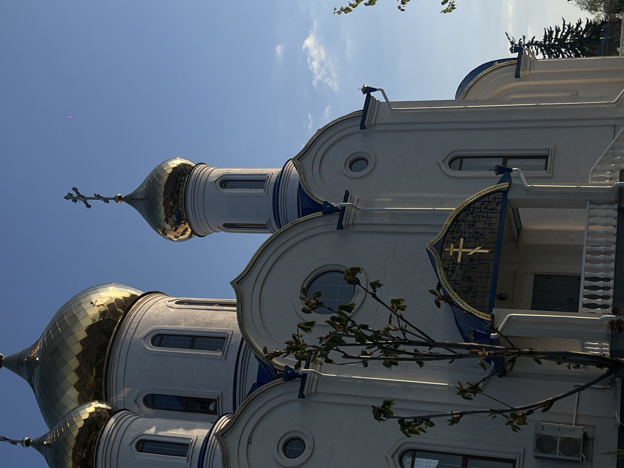 This vertical image depicts an ornate Orthodox church, likely located in Novopokrovskaya, Russia, under a clear blue sky with sparse white clouds. The main subject is the church building, characterized by its bright white walls contrasted with deep blue accents and intricate gold detailing.
The church features multiple golden, onion-shaped domes, several of which are visible. The largest and tallest dome, topped with a distinctive Orthodox cross, is prominent on the left side of the frame. Another smaller gold cross is integrated into a decorative blue and gold metal panel above an entrance, which also features white balustrades.
Architectural details include various window shapes – arched, rectangular, and small circular windows – set within the white facade. Gold trim and decorative elements adorn the edges of the roofs and architectural contours. Parts of a tree with green leaves partially obscure the building in the foreground, adding a natural element to the scene.
The bright sunlight suggests either morning or late afternoon, illuminating the golden domes and creating clear contrast between the white walls and blue details. No people or specific events are visible, presenting a serene view of the architecture. A small, indistinct pink speck is noticeable in the sky near the main dome. An outdoor air conditioning unit is visible on the lower right side of the building. No discernible text is present beyond the religious symbolism of the crosses.