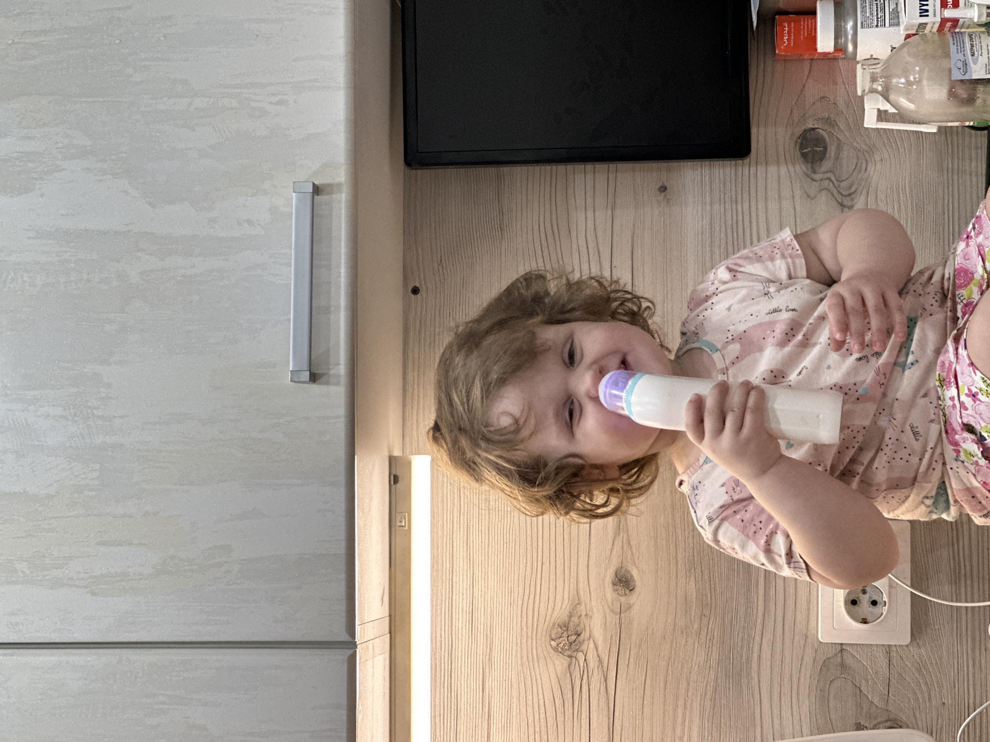 A child is shown from an overhead perspective, laying on a wood-grained surface, with their head toward the left. The child is holding a white bottle with a purple nipple. To the left of the child is a light-colored cabinet with a silver handle, and a black rectangular object is positioned above the child. To the right of the child are various medicine bottles and other objects. An electrical outlet is visible near the child's body. The location is Chaikovsky, Russia.