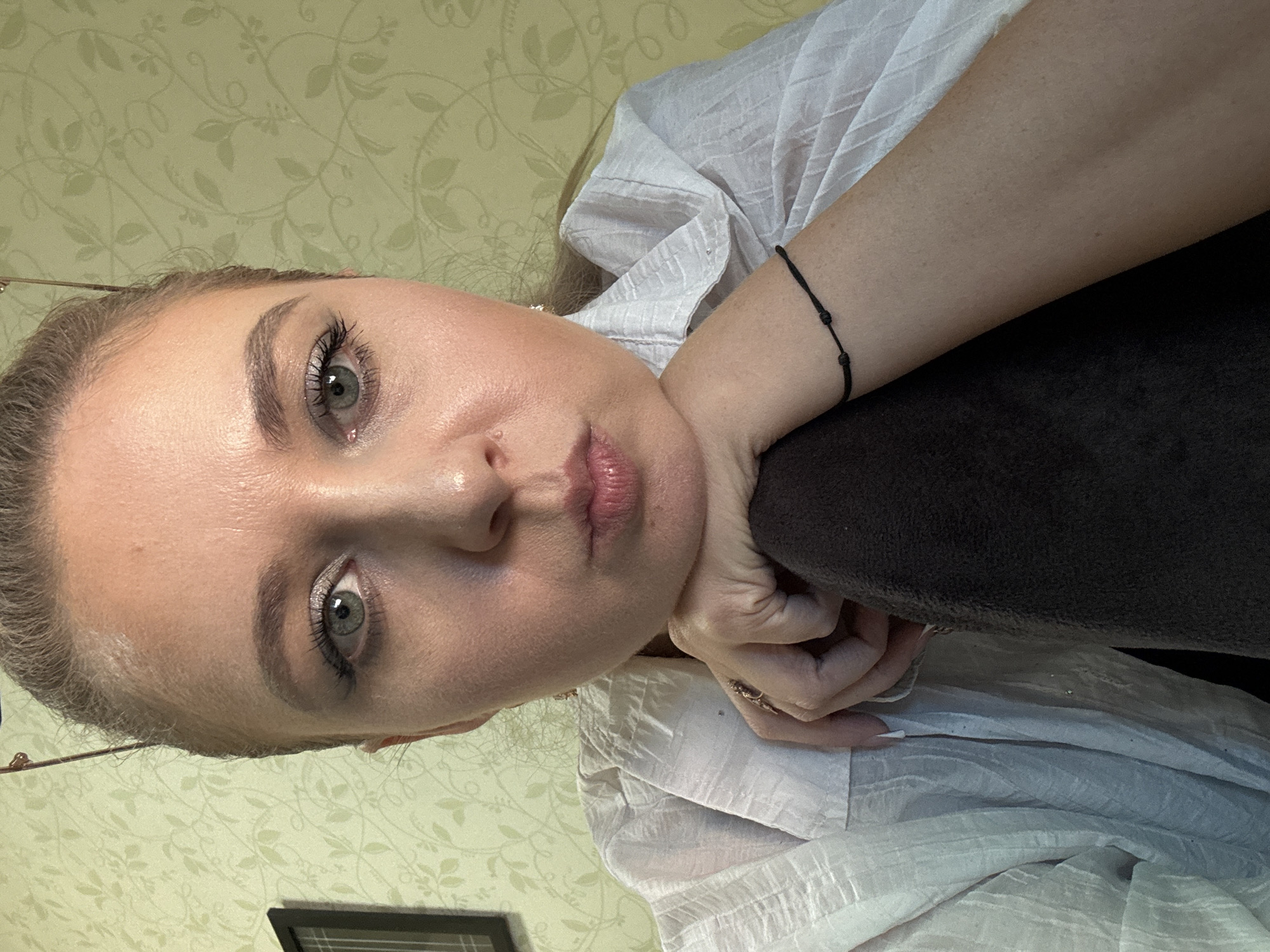 The image is a close-up, selfie-style photograph of a woman's face and upper body. She has light skin, blonde hair pulled back, and green eyes. Her lips are slightly parted, and her chin rests on her left arm. She is wearing a white, textured button-down shirt. A black string bracelet with three knots is visible on her right wrist. Her left hand is visible, resting on her arm, with a ring on her index finger. The background appears to be a wall with a green, floral wallpaper pattern. To the left of the woman, a dark framed picture is partially visible. The lighting is bright, illuminating the woman's face and highlighting the texture of her shirt and the wallpaper. The overall impression is of an indoor setting.