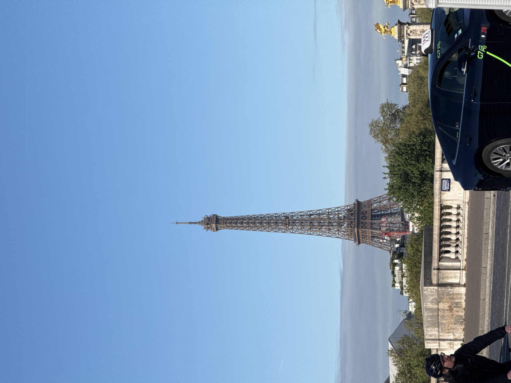 The image shows the Eiffel Tower in Paris, France, under a clear blue sky. The tower is the central subject, seen from a ground-level perspective, likely from a bridge. A dark-colored car, marked as a "G7 Taxi" with a green line, is visible on the right side of the frame, along with a cyclist wearing a helmet and jacket. In the background, a glimpse of golden statues, trees, and Parisian buildings can be seen, suggesting the location is near the Place de la Concorde. A concrete bridge with a stone balustrade is visible, indicating the scene is set alongside the Seine River.