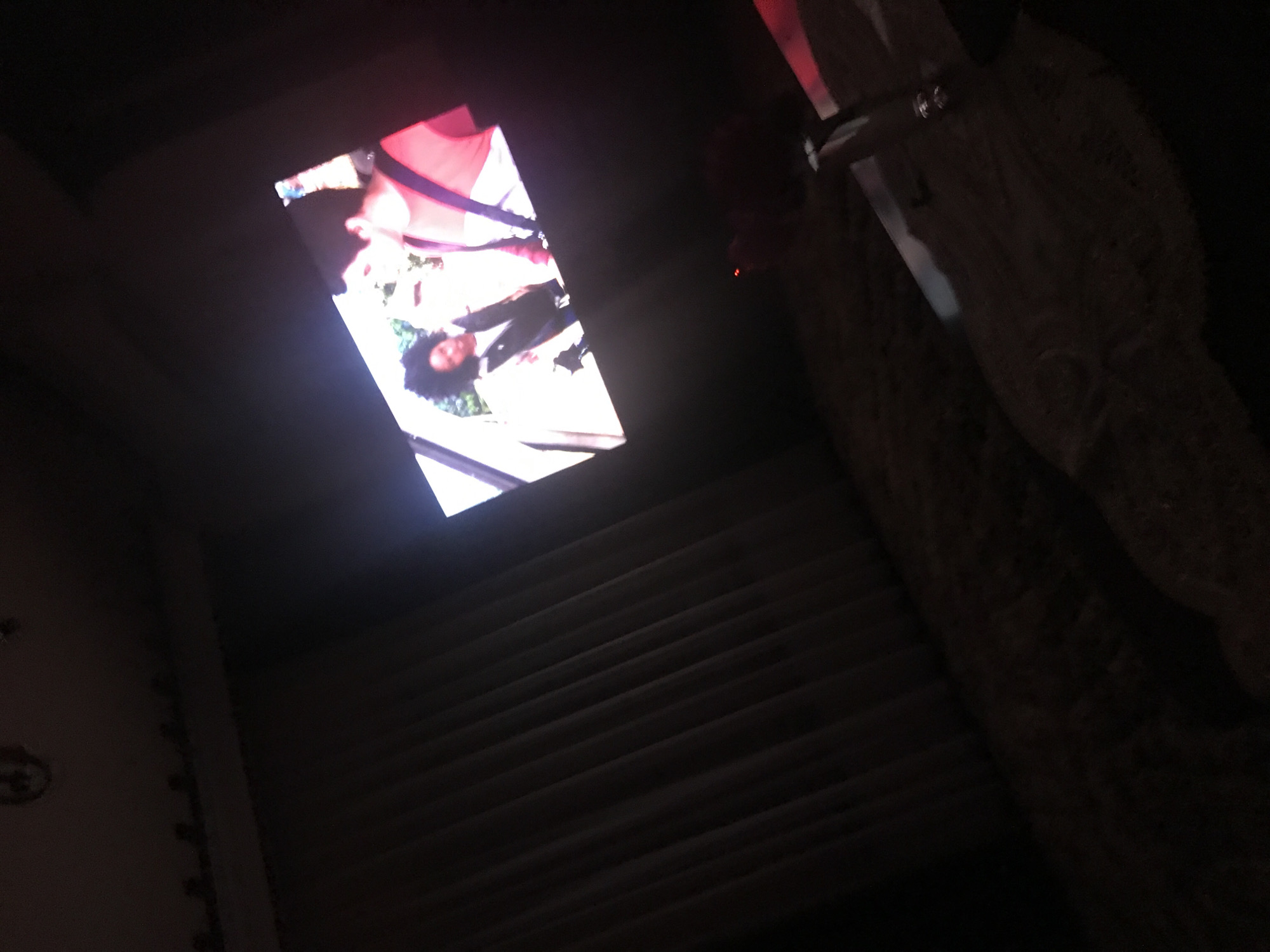 The image shows a low-angle view of stairs within a dimly lit space, likely indoors. In the upper left, a rectangular screen displays a brightly lit scene featuring a woman with dark, curly hair wearing a white top and a pink garment. In the upper right corner, there is a partially visible stone wall with what appears to be a metallic object attached to it. The city and location within Yerevan, Armenia, cannot be determined from the visual data.