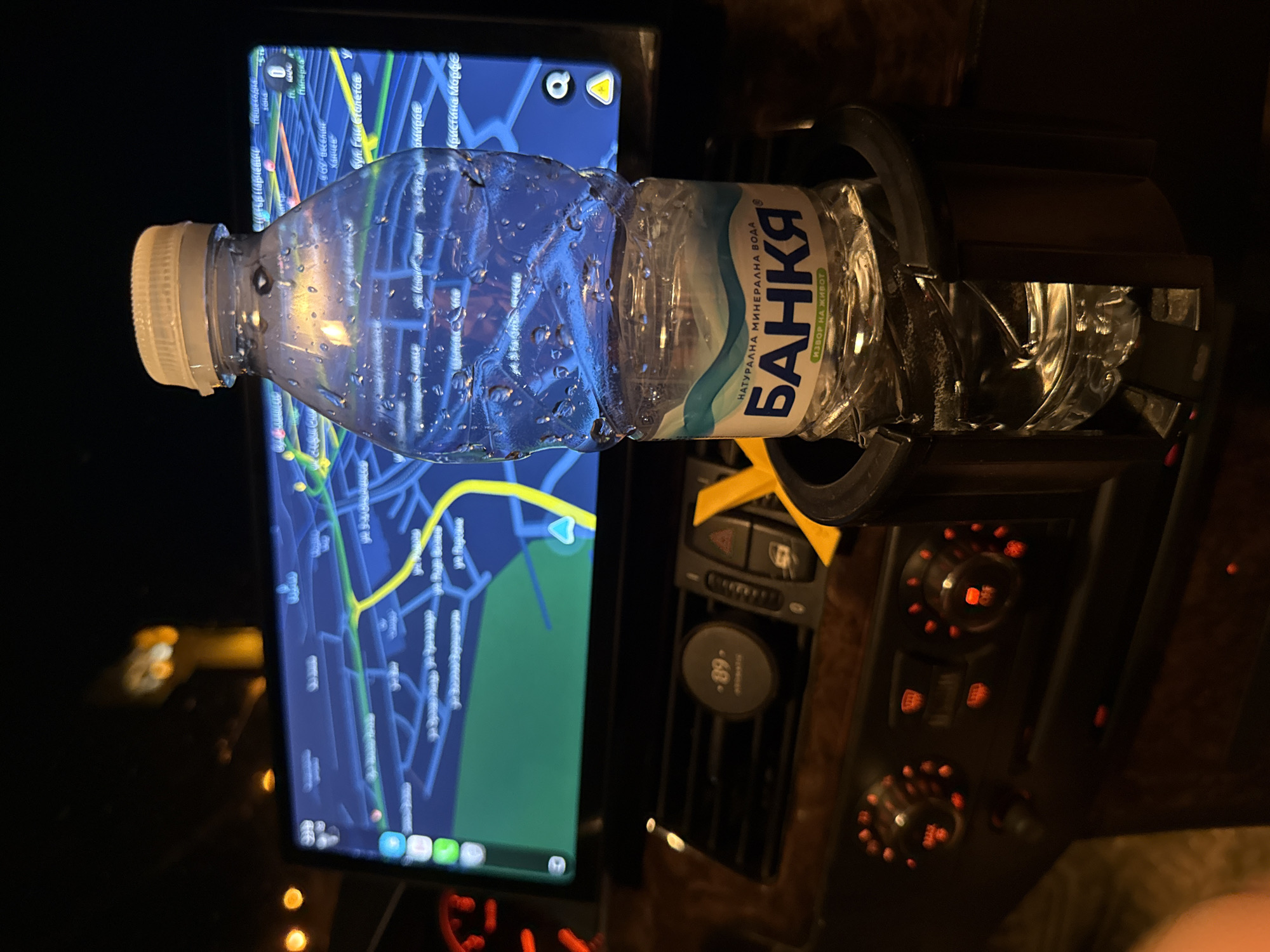 The image captures an interior view of a vehicle at night, with a focus on a partially full bottle of "Bankya" mineral water placed in a cup holder. The water bottle has condensation on its exterior, indicating it is cold. Behind the bottle, a large car infotainment screen displays a navigation map, seemingly of a city, with roads and points of interest visible. The map interface suggests a location within Bulgaria, as indicated by Cyrillic script for place names. The vehicle's dashboard is visible, featuring wooden trim, climate control knobs with red illuminated indicators, and an air vent with a small circular device attached. The ambient lighting from the car's interior and the screen creates a dimly lit environment. The overall scene suggests the vehicle is stationary, possibly parked, or driving at night in an urban setting.