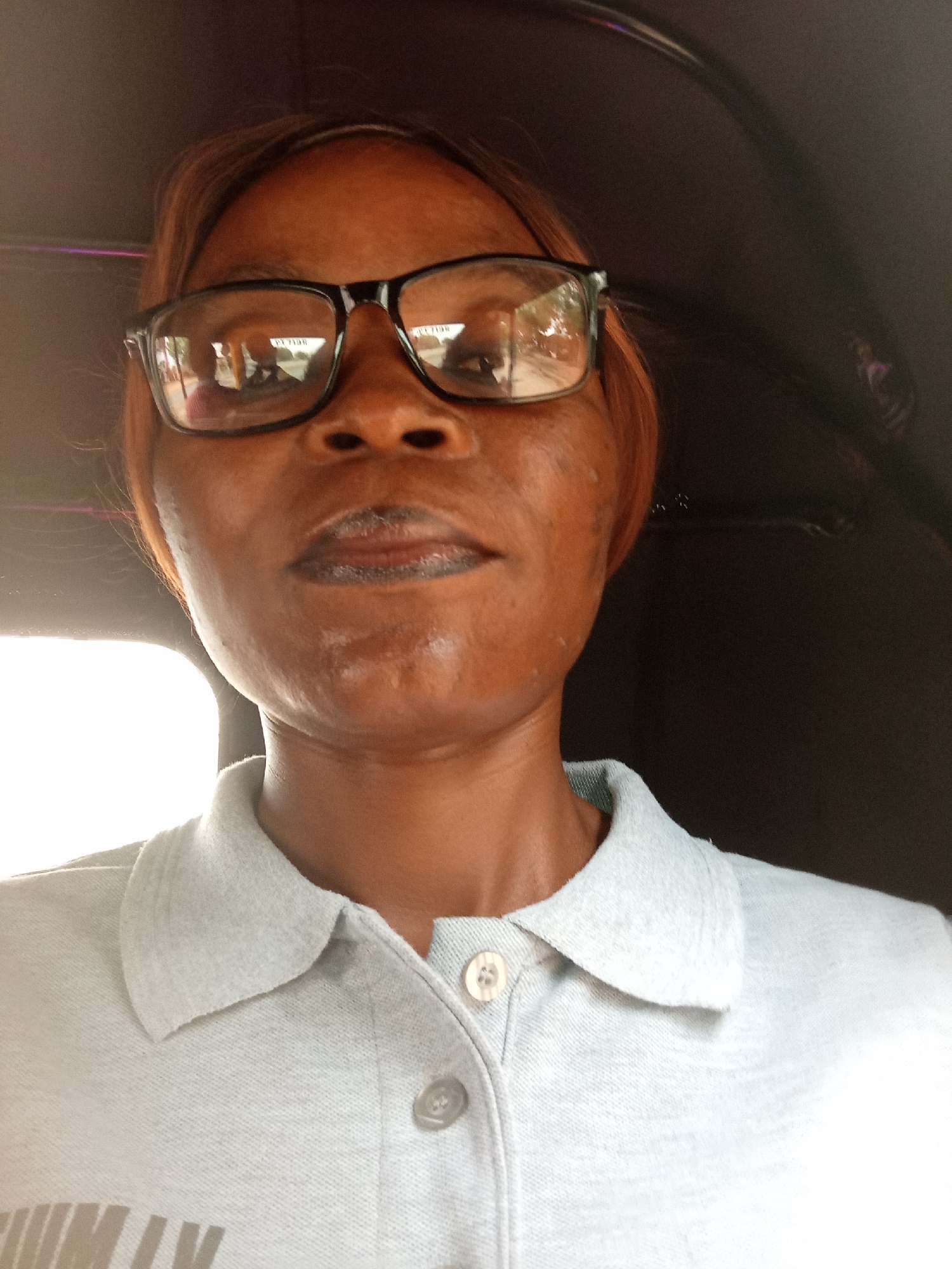 A close-up, waist-up selfie captures a woman wearing glasses and a light blue collared shirt. The woman's face is centered in the frame, her expression neutral. She has dark skin and reddish-brown hair, which is pulled back. The shirt appears to be a polo shirt with buttons. The background is dark and slightly out of focus, suggesting the photo was taken inside a vehicle, likely an auto-rickshaw, given the shape of the overhead canopy. The lighting is bright, with a significant light source to the left of the frame, casting shadows and highlighting the woman's skin texture and the fabric of her shirt. The scene appears to be in Zanzi, Nigeria.