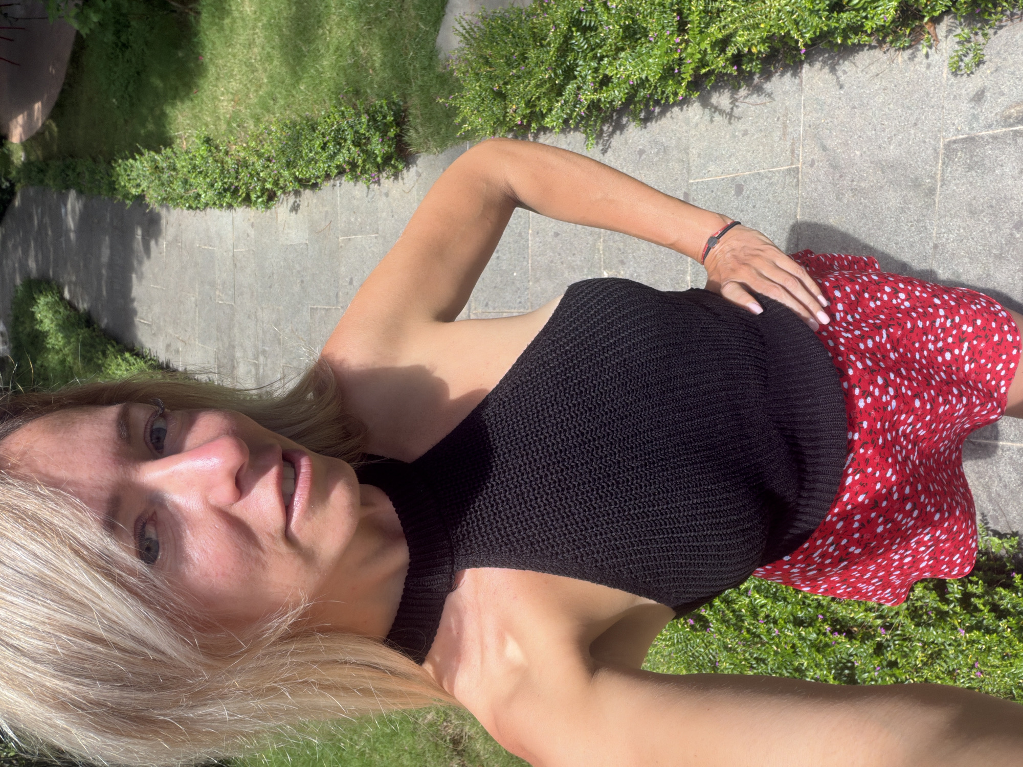 In the city of Xuân Trường - Đà Lạt, Vietnam, a woman with blonde hair is captured in a selfie taken outdoors. She is wearing a black halter-neck top and a red skirt with white polka dots. The woman is standing on a paved walkway lined with lush green bushes. The walkway appears to be made of gray stone slabs, and sunlight casts shadows on its surface. The background consists of more greenery and hints of architectural elements, suggesting a well-maintained garden or park setting. The woman's pose is casual, with one hand on her hip. The image captures a moment of outdoor leisure in a scenic environment.