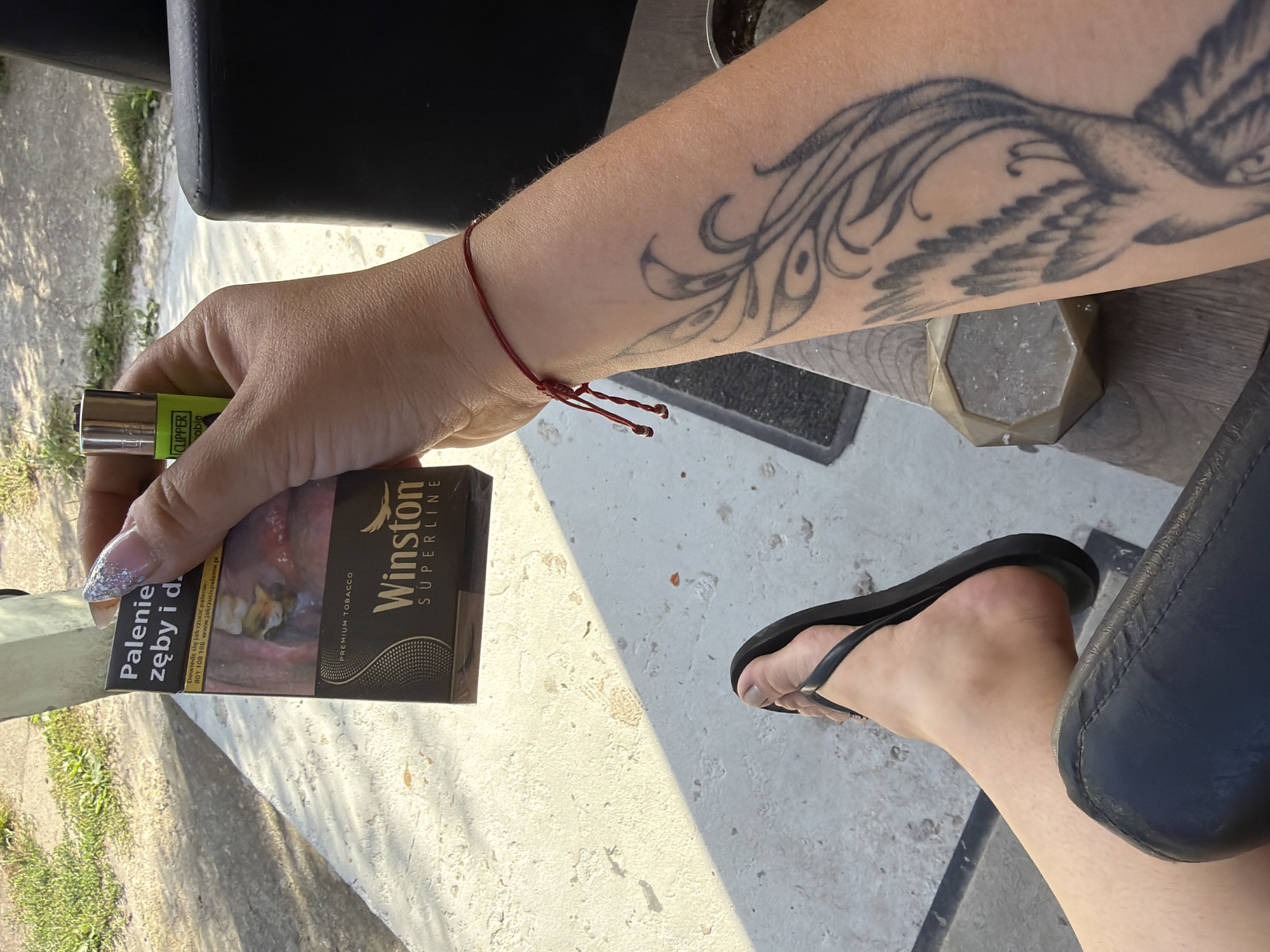 A close-up, high-angle shot features a person's hand holding a pack of Winston Superline cigarettes and a green Clipper lighter. The pack displays a graphic warning about smoking's effects on teeth and gums, along with Polish text. The hand has long, decorated fingernails. A thin red string bracelet is tied around the wrist, above a tattoo of a bird or feather design on the forearm. In the foreground, a bare foot wearing a black flip-flop is visible, resting on a black surface, likely a chair. The background shows a light-colored, rough-textured ground, possibly concrete, with some greenery and shadows suggesting an outdoor setting. The scene appears to be in Szczecin, Poland, judging by the Polish text on the cigarette pack, but no specific landmarks or identifiable locations within the city are visible.