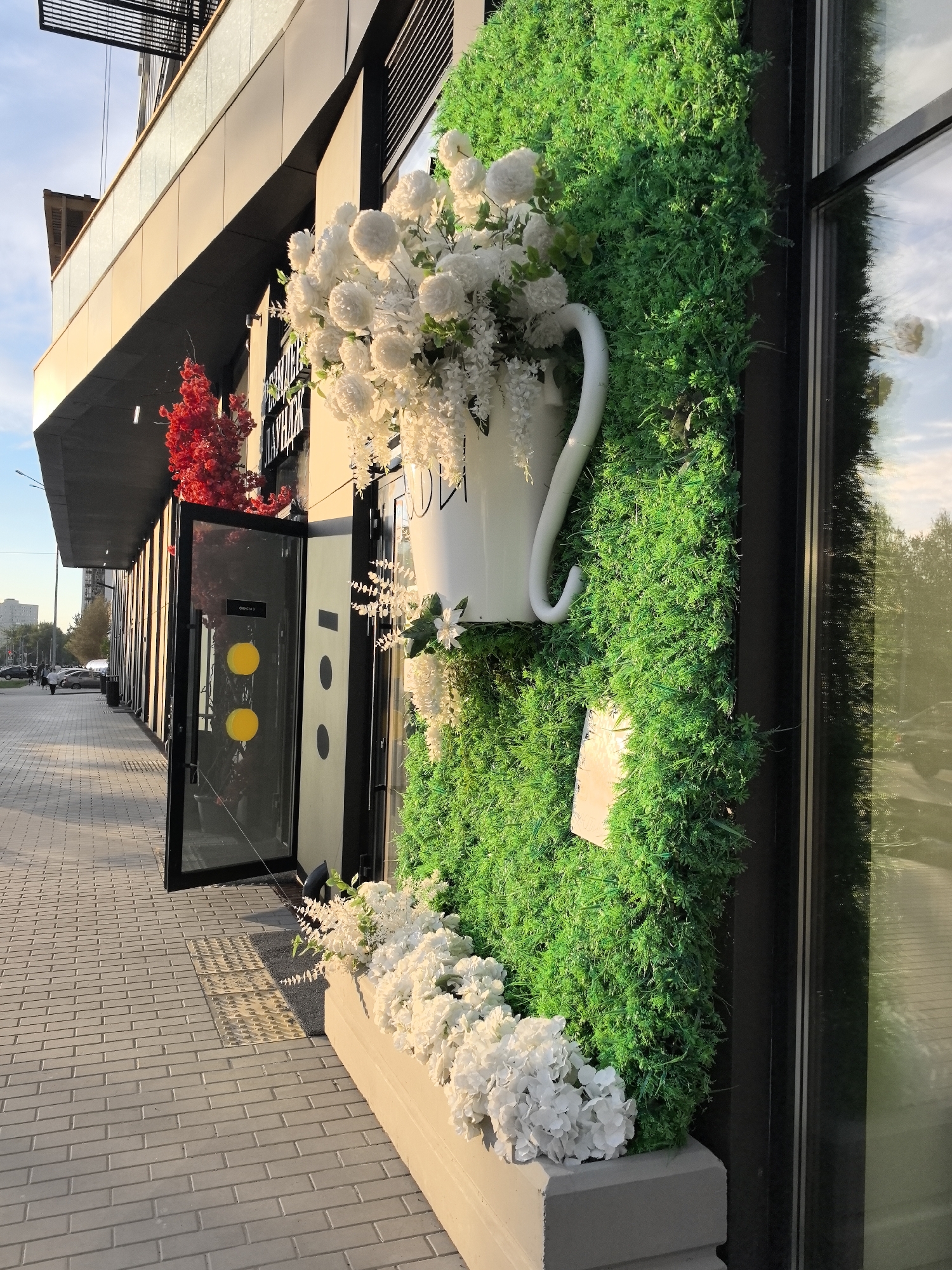 This is a photograph of a building in Perm, Russia. The image features a modern building facade with a distinctive display of artificial greenery. A large, white decorative pitcher, filled with white flowers, is mounted on a wall covered in faux foliage. Below the pitcher, a planter box also overflows with matching white flowers. The building's architecture includes dark window frames and a glass door with yellow circular details, possibly indicating the entrance to a commercial establishment. The sidewalk is paved with gray bricks. The lighting suggests it's daytime, with some sunlight reflecting off the glass surfaces. In the background, there's a street with cars and distant buildings, indicating an urban setting.