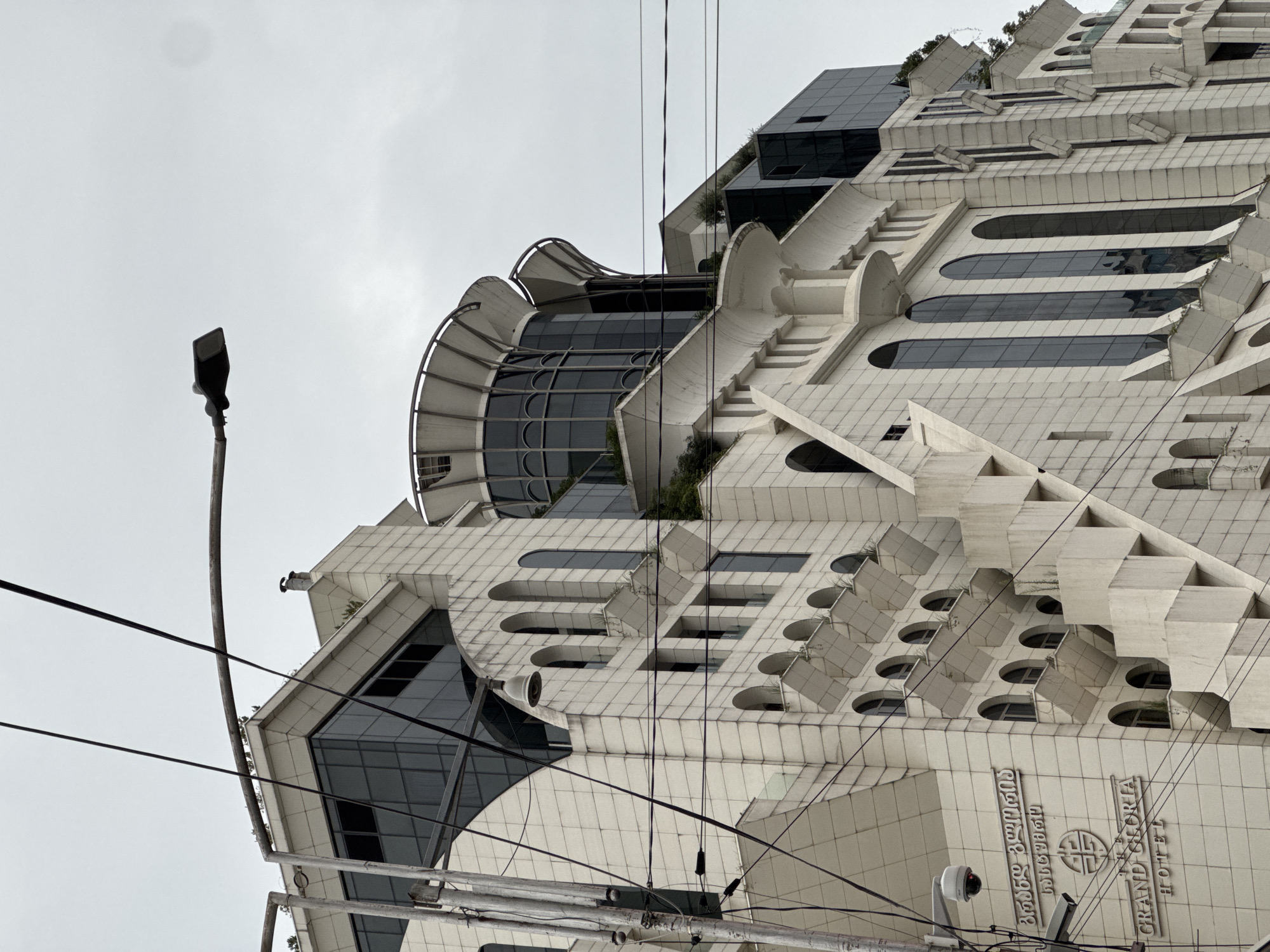 The image shows the Grand Gloria Hotel in Batumi, Georgia. The hotel is a modern building with a white facade and numerous windows. The building has a unique architectural style with several protruding balconies and geometric shapes. There are also power lines and a lamppost visible in the foreground. The sky is overcast, and the overall impression is one of a cloudy day. The text on the building is in Georgian and English, identifying it as "THE GRAND GLORIA HOTEL". Two CCTV cameras are also visible on the left side of the building.