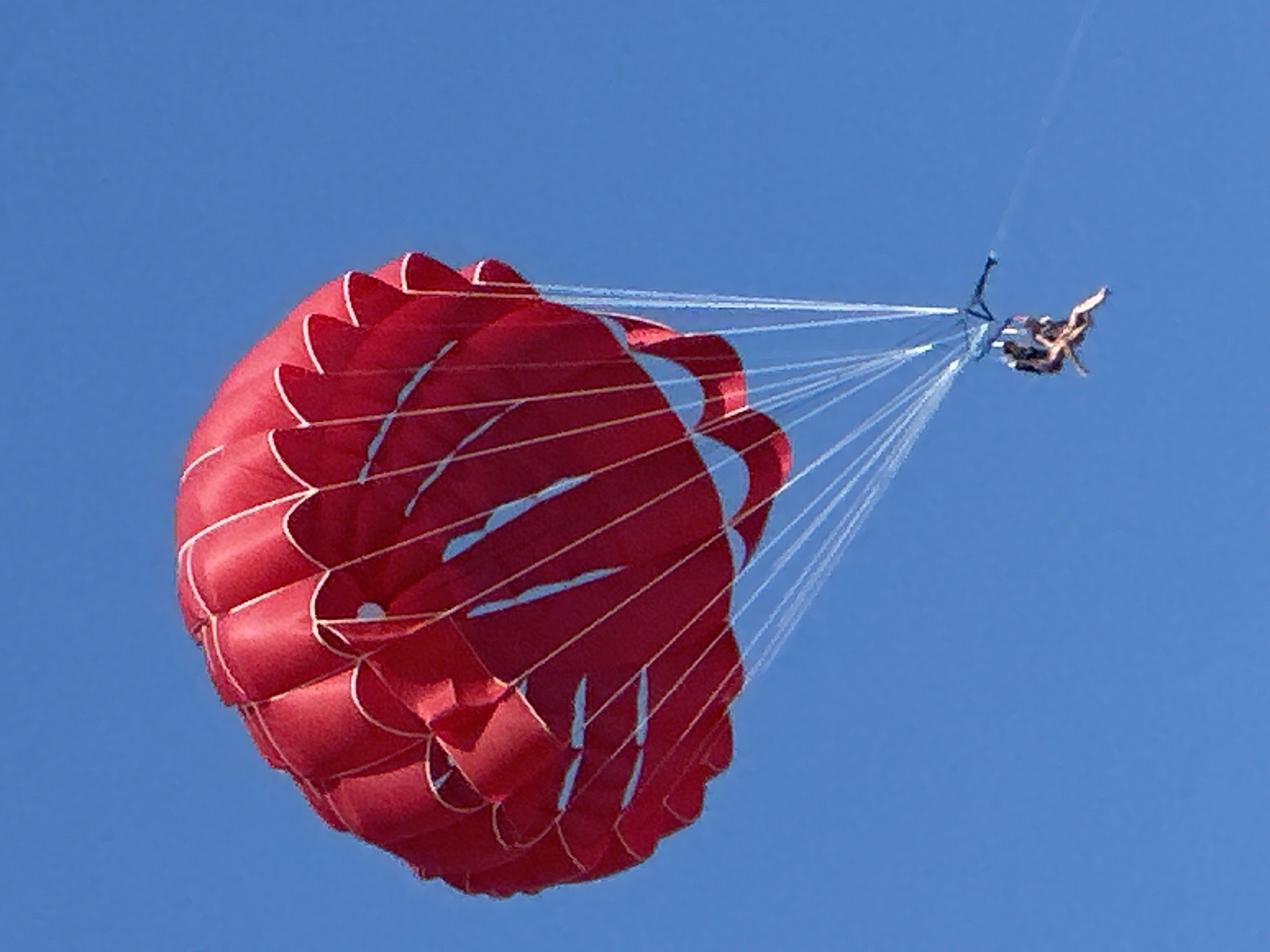A bright red parachute, filled with wind, floats against a clear blue sky. The parachute is suspended by thin white lines, which converge at a point below, where a person is attached. The person appears to be parasailing, with their legs dangling and arms outstretched. The parachute is large and circular, with distinct sections and white trim along the edges. The image is taken from a low angle, looking up at the parachute and the person against the vast expanse of the sky. The scene suggests an activity taking place in an open area, likely over water given the common association with parasailing. There is no indication of the city of Boreti, Montenegro, in the image itself.