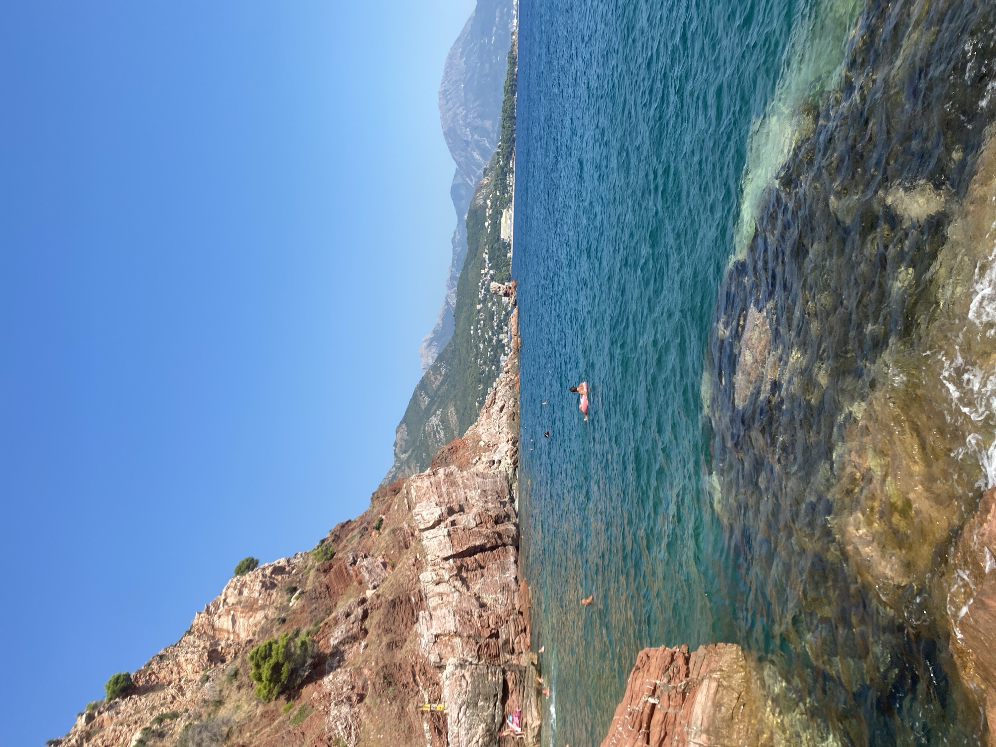 The image depicts a coastal scene with clear turquoise water and a rocky shoreline. In the foreground, reddish-brown rocks are partially submerged in the water, with some areas revealing the sandy or rocky seabed below the surface. The water is calm, with gentle ripples.
Further out in the water, several individuals are visible. One person is swimming or floating on an inflatable ring. Others appear to be swimming or wading closer to the shore.
To the left, a steep, rocky cliff face rises from the water. This cliff is characterized by layered reddish-brown rock and sparse green vegetation growing from crevices. A small, yellow platform structure is visible on the cliff face near the water's edge, possibly indicating a diving board or marker.
In the background, a range of mountains stretches across the horizon, covered in green vegetation. A coastal town or village is nestled along the lower slopes of these mountains, with buildings and roads visible. The sky is a clear, bright blue with no clouds. The overall impression is a sunny day at a Mediterranean beach or cove. This scene is consistent with locations along the coast of Sutomore, Montenegro, known for its rocky shores and mountainous backdrop.