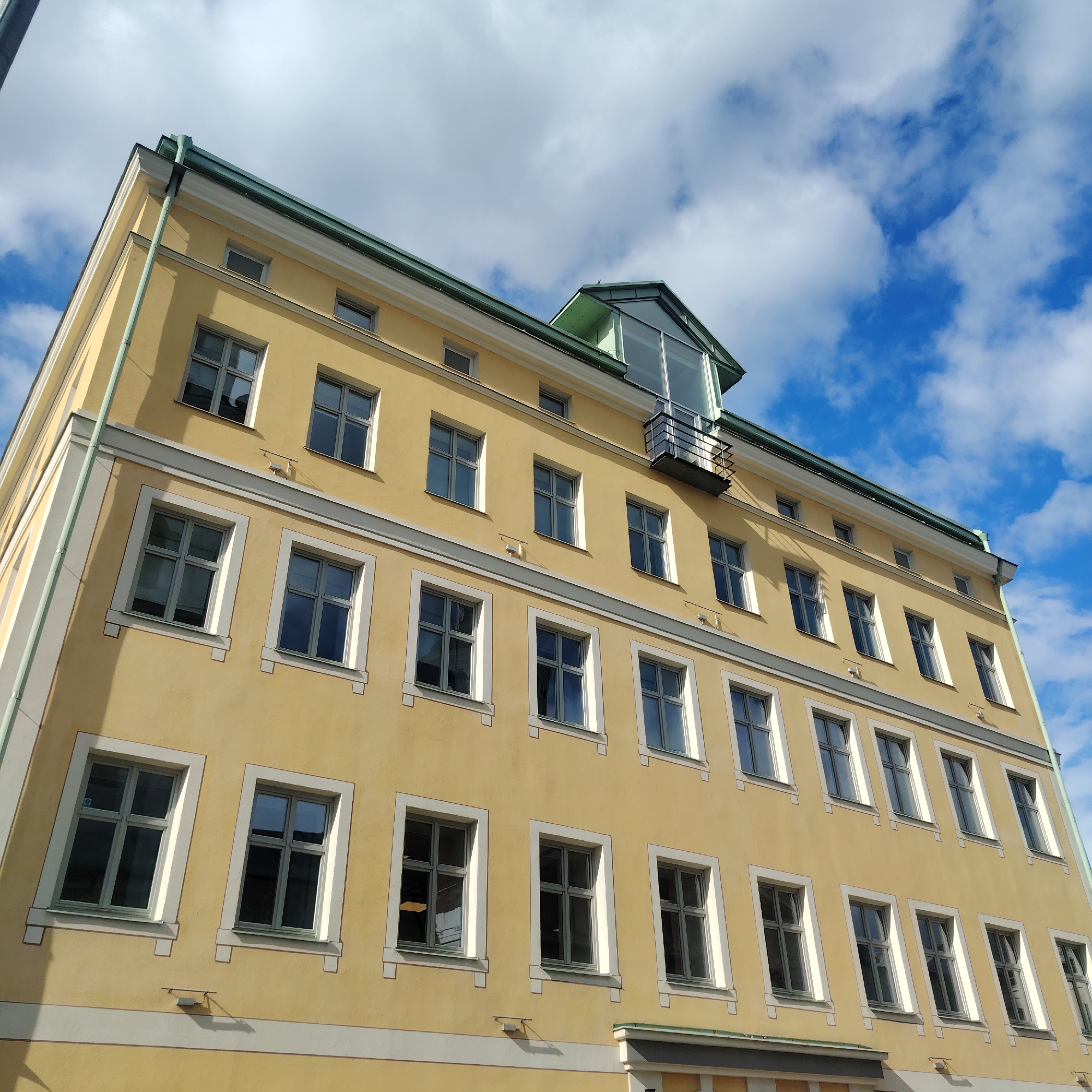 The image shows a yellow building with numerous windows in Riga, Latvia. The building has four visible stories and an additional glass-enclosed structure on the roof with a small balcony. The facade is a muted yellow with white trim around the windows and a grey stripe separating the first floor from the rest. Above the top floor windows is a thin green roof and a white line along the top. The sky is blue with white clouds. No people are visible in the image.