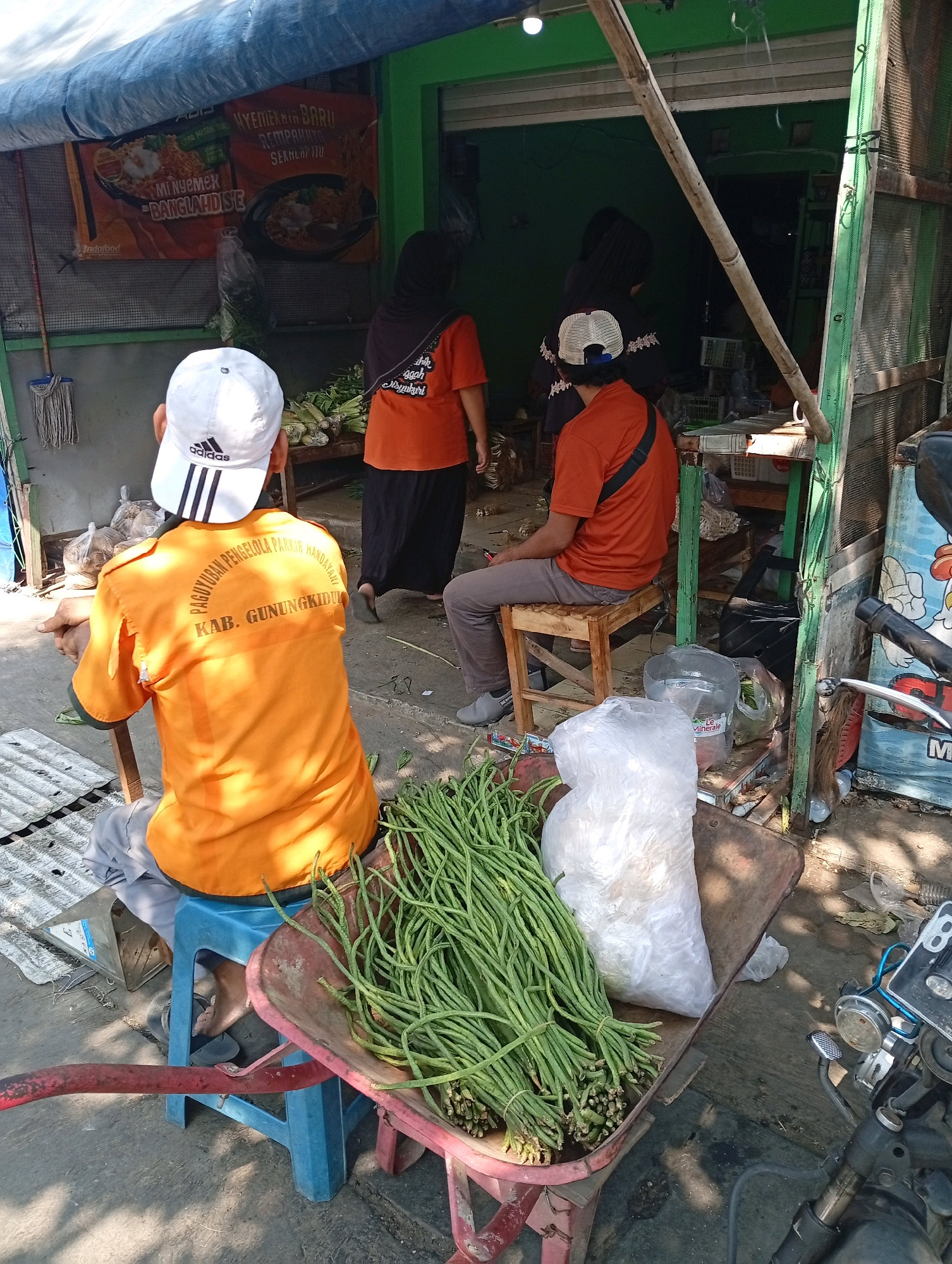 The image depicts an outdoor scene in Playen, Indonesia, likely a roadside stall or market area. In the foreground, a man wearing an orange shirt with the text "PAGUYUBAN PENGELOLA PARKIR KAB. GUNUNGKIDUL" and a white Adidas cap sits on a blue plastic stool. In front of him, a red wheelbarrow holds a large bunch of green long beans and a white plastic bag. To the right, a motorcycle is partially visible.
In the background, two women are standing near an open storefront with a green facade. One woman wears a dark hijab and an orange shirt, while the other wears a dark hijab and an orange shirt as well. A banner with Indonesian text and images of food is hanging on the wall behind them. Various goods and structures, including wooden beams, metal mesh, and a blue tarp, are visible, suggesting a functional, possibly informal, commercial setting. The ground is paved, and sunlight creates shadows, indicating it is daytime.