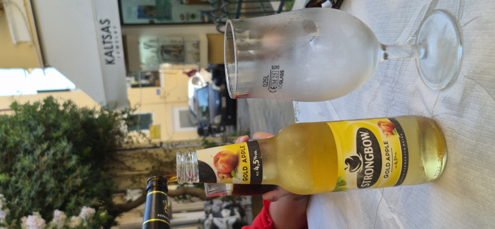 In Corfu, Greece, an outdoor cafe scene unfolds with a focus on beverages. A bottle of Strongbow Gold Apple cider is being poured into a clear glass filled with ice, positioned on a white, textured tablecloth. To the left of the bottle, a second glass, partially filled with a dark liquid, is visible. In the background, a street scene features buildings, a tree with lush green foliage, and parked cars. A jewelry store named "KALTSAS JEWELRY" is prominently displayed on one of the buildings. The overall ambiance suggests a relaxed, daytime setting in a Mediterranean locale.