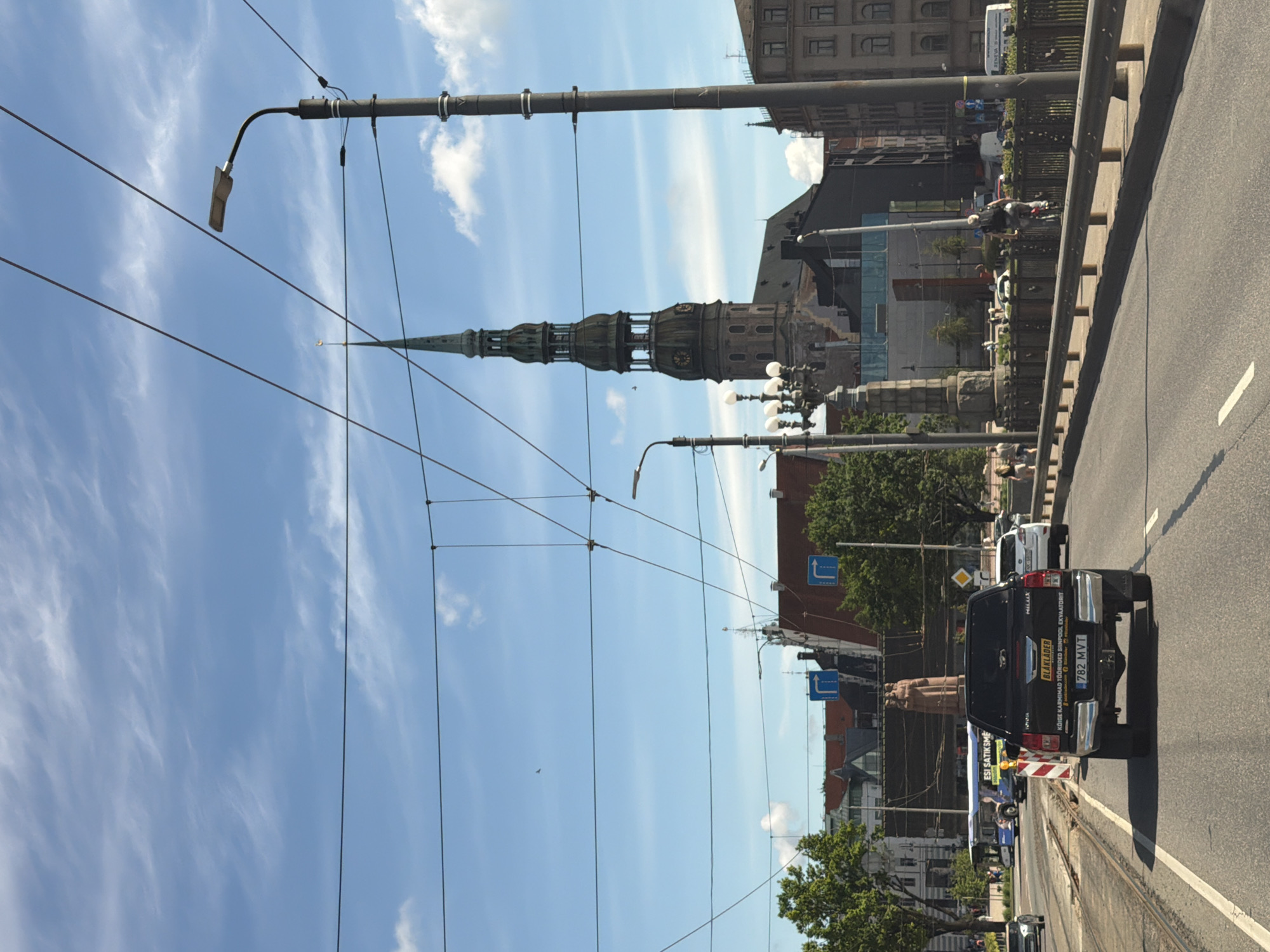 The image depicts a street view in Riga, Latvia, featuring the spire of St. Peter's Church prominently in the background. In the foreground, a black Toyota Hilux pickup truck with a yellow license plate is driving on a road. The truck has text on its tailgate, including "BLAKLÄDER" and a Latvian license plate "782 MVX". A bus with "ESI SATIKŠIES" written on it is visible further down the road, along with other vehicles and pedestrians. The scene includes overhead wires, streetlights, and tram tracks, suggesting an urban environment. The weather appears to be sunny with some clouds in the sky.