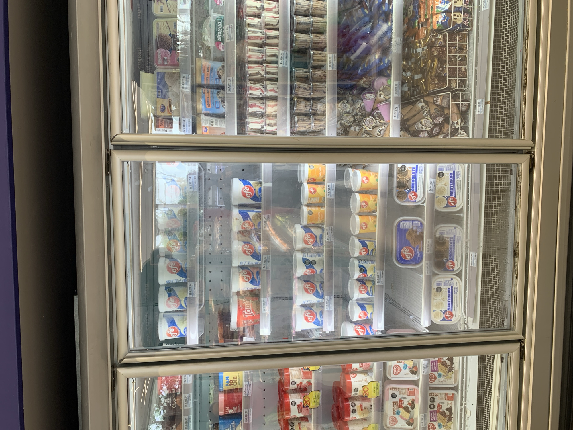 This image presents a detailed view of a three-door commercial freezer unit, likely situated within a supermarket or convenience store in La Asuncion, Venezuela. The freezers are predominantly stocked with various frozen desserts, primarily ice cream products from the brand "efe," identifiable by its distinctive red and blue logo.
The leftmost freezer section, partially visible, displays multi-pack ice cream tubs and boxes on its upper shelves, with prices in Venezuelan Bolivars (Bs.) such as "Bs. 3,232.54." Lower shelves in this section contain rectangular plastic containers of ice cream, some pink and some white, also featuring the "efe" brand, alongside smaller red and white tubs marked with "20" and prices around "Bs. 2,223.91" and "Bs. 3,223.91."
The central freezer unit is neatly organized with multiple shelves. The top shelf holds white tubs with blue lids and the "efe" logo, priced at approximately "Bs. 3,101.24." Below this, a red tub labeled "TODDY" is visible amongst more "efe" brand tubs. Subsequent shelves showcase various sizes of "efe" branded white tubs with blue lids, including those labeled "MANTEGADO" (likely vanilla) and "CHOCOLATE," with prices ranging from "Bs. 2,684.77" to "Bs. 3,943.57."
The rightmost freezer unit features a diverse array of frozen goods. Its upper shelves contain a mix of rectangular ice cream tubs and cups from various brands, including "CRAQ," "MANTECADO," and "COOKIES AND CRE," with prices up to "Bs. 4,925.59." The middle shelves are densely packed with numerous small, clear plastic cups containing layered desserts, stacked in rows, each priced at "Bs. 509.31." The lower shelves and wire baskets are filled with individually wrapped ice cream novelties, including cones (some labeled "SUPER CONO") and bars, with prices like "Bs. 525.73" and "Bs. 788.49."
The freezer doors are made of glass, reflecting overhead lighting and a blurry, indistinct background. The metal frames of the freezers show some wear, and there is visible condensation and a brownish discoloration along the bottom seals of the freezer doors, suggesting age or a need for cleaning. No individuals are present or engaged in any activity within the visible frame of the image. The environment suggests an active retail setting, with products clearly priced for sale.