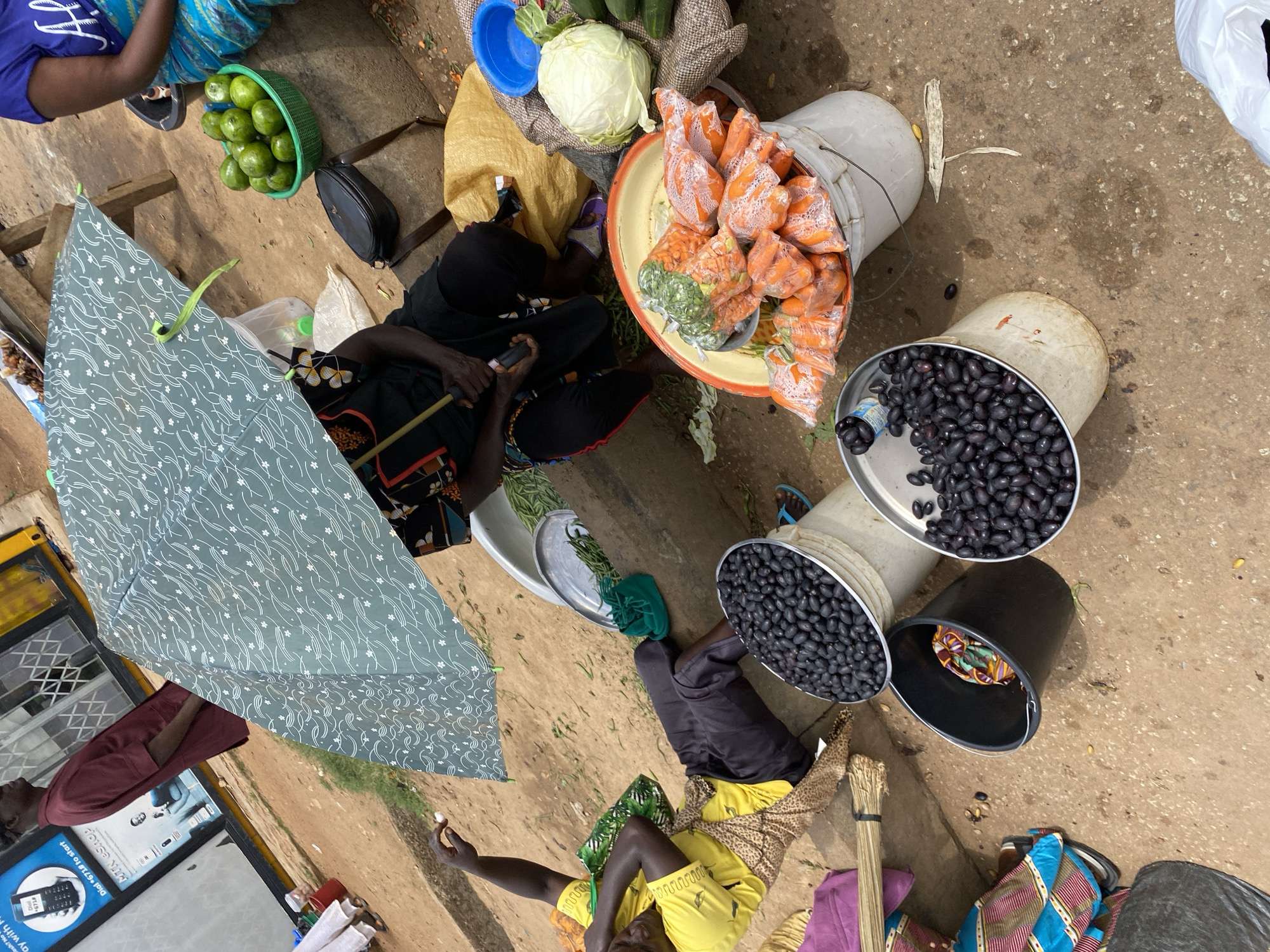 In the city of Bukuru, Nigeria, a street market scene unfolds under a patterned green umbrella. Two women are present. One, partially obscured by the umbrella, wears a black top and dark trousers, her hands holding an umbrella pole and a small object. The other woman, wearing a yellow t-shirt and dark trousers, reclines on the ground, her arm raised and hand holding a small white object. She is positioned next to a large white bucket filled with dark, oval-shaped fruits, likely olives.
In the foreground, a black bucket containing colorful fabric sits on the ground. To the right, another white bucket overflows with pre-packaged carrots, their bright orange color visible through the plastic. Behind this, a large cabbage rests on a textured surface, next to a blue plastic basin and a green basket containing more green, round fruits, likely limes or small oranges. A person in a blue t-shirt and blue patterned fabric is visible to the upper right, seated near the green basket. The ground is a mixture of sand and dirt. In the background, a man in a maroon tunic stands near a shop window with an advertisement for "MTN eShop." The overall atmosphere suggests a bustling, outdoor market environment.