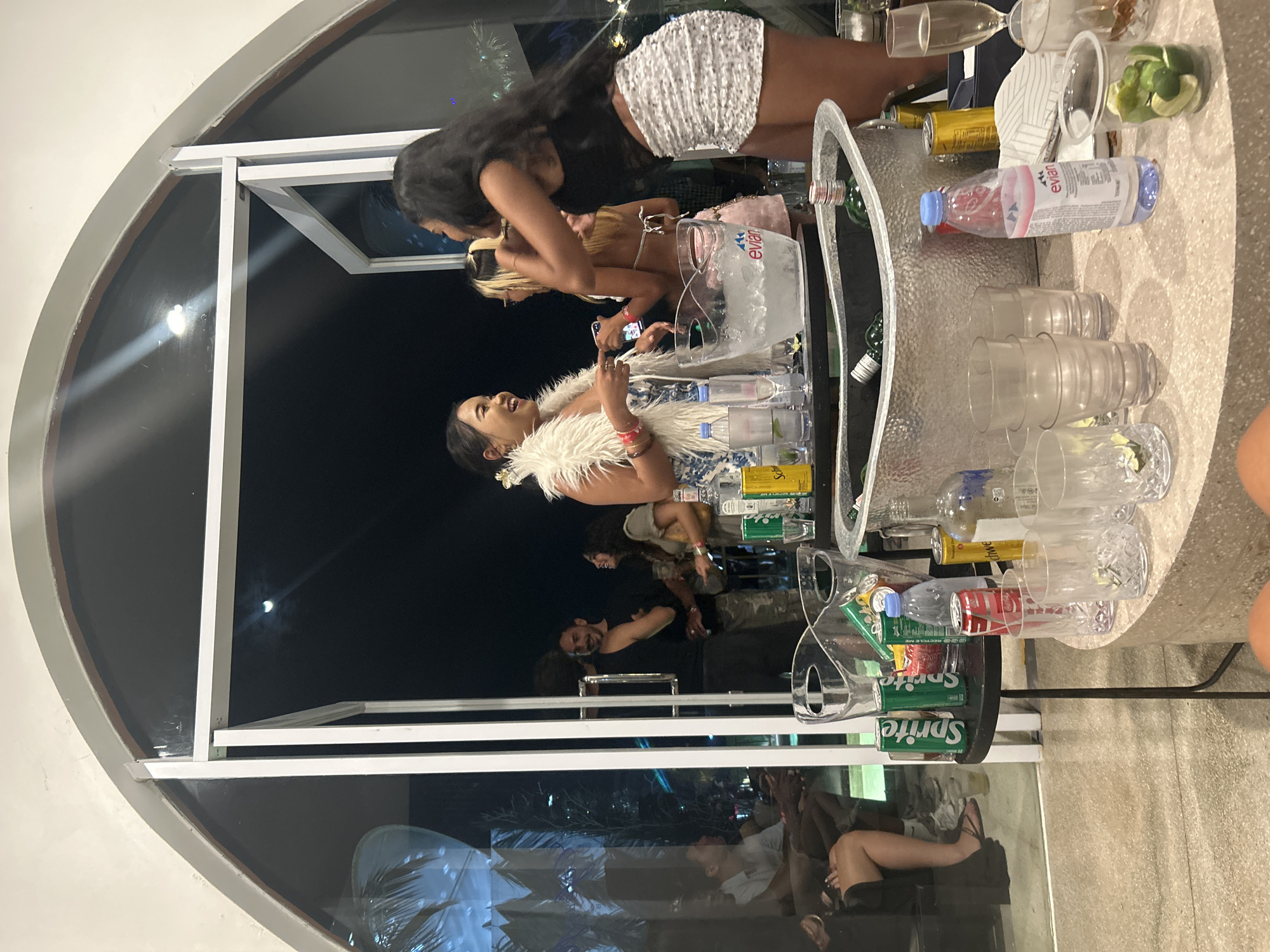 The image depicts a nighttime social gathering at a venue in North Kuta, Indonesia, characterized by an indoor-outdoor setting with large glass doors. In the foreground, a round, speckled counter displays various beverages and drinking glasses. Prominently featured are two large galvanized tubs filled with ice and bottles, one labeled "Evian." Several glass tumblers, some containing lime wedges, are arranged on the counter, alongside cans of Sprite and Coca-Cola. A bottle of Evian water stands upright on the counter.
Through the glass doors, several individuals are visible. Two women in the center, dressed in party attire, appear to be interacting, with one wearing a white fur stole and the other a sequined skirt. Behind them, another woman is seen in a black top and a sequined skirt, facing away from the camera. Further in the background, a man in a black tank top is seen laughing. Other guests are seated and standing within the venue. The outdoor area visible through the doors appears to be illuminated with decorative lights, and palm fronds suggest a tropical environment. The overall scene conveys a lively atmosphere of a party or social event.