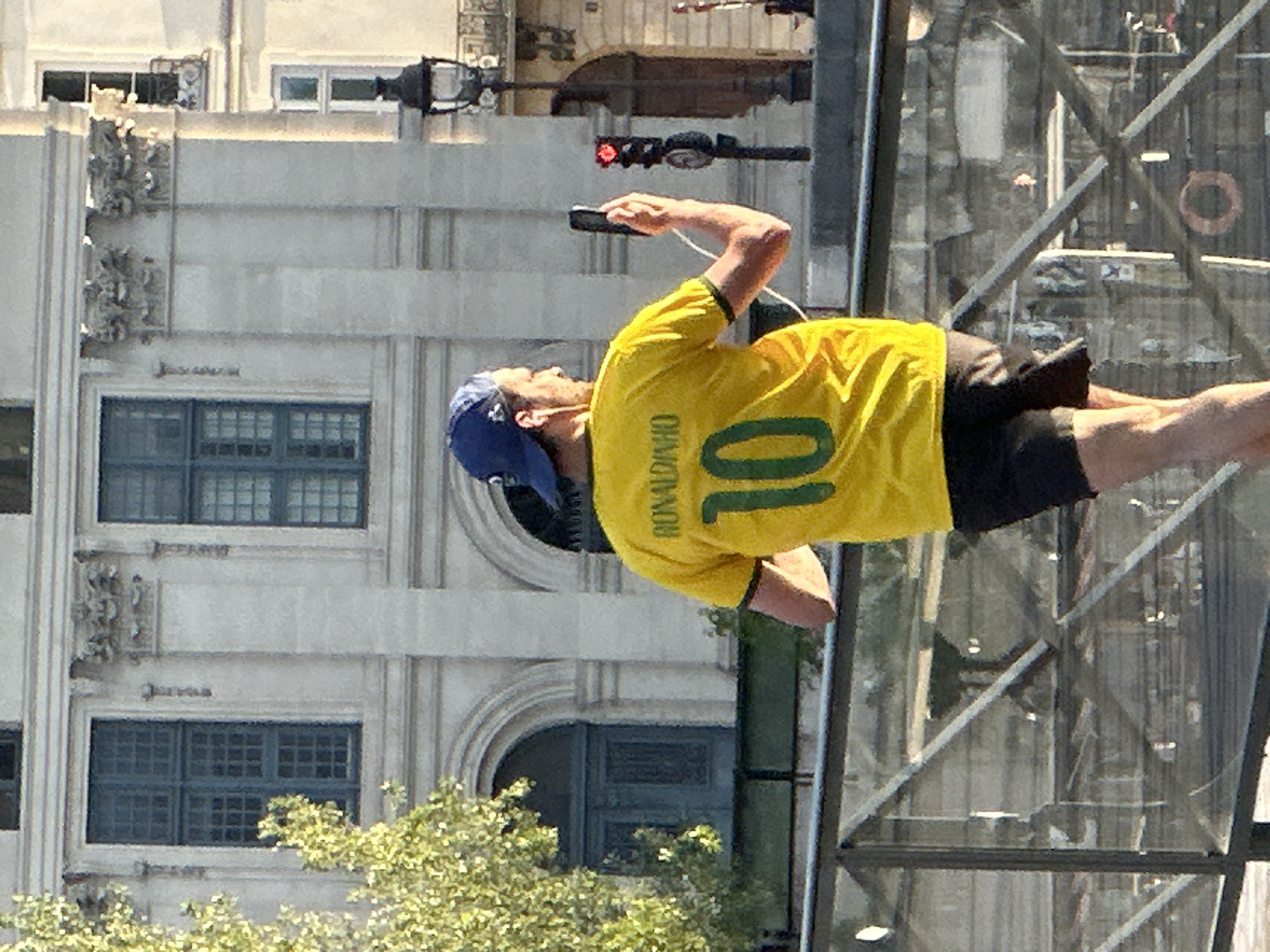 A man wearing a yellow Brazil soccer jersey with the name "Ronaldinho" and the number "10" on the back is seen using his phone in front of the Louvre Museum in Paris, France. He is wearing a blue cap and black shorts. The Louvre's glass pyramid structure is visible in the background, along with a building and a tree. A street lamp with a red light is also present.