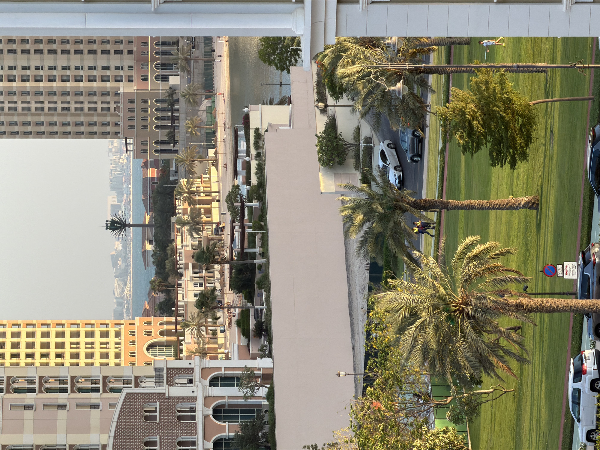 This high-angle, vertical outdoor photograph depicts a coastal urban landscape during the late afternoon, characterized by warm, golden lighting. The scene is set in an unidentified city.
In the foreground, a meticulously maintained green park spans the lower half of the image. It features numerous mature palm trees with visible trunks and lush fronds, alongside several smaller deciduous trees. An asphalt road with white markings runs through the park. A white SUV is parked on the left, a grey SUV in the center, and a white sedan is partially visible on the far right. Two individuals in high-visibility vests are visible near the white SUV on the left, while a person in a light-colored dress walks on the grass to the far right. Several ornate black lampposts illuminate the park, and a red circular "No Entry" sign with a secondary sign displaying text in Arabic and English ("No Waiting") is visible near the center.
The midground is dominated by architectural structures. On the left, a multi-story building with a light-toned, elaborate facade features multiple arched windows, balconies, and decorative patterns on its lower levels. On the right, a taller, more uniform multi-story building with a beige facade and numerous rectangular windows stands prominently. A portion of a white classical column is visible on the extreme right edge. Between these buildings, a long, pale peach-colored concrete wall forms the base of a terraced area, above which landscaped gardens with trees and shrubs are visible. Further back, a sandy beach is dotted with several dark sun umbrellas and approximately a dozen small figures, some seated, others walking. A calm, light blue-green body of water (likely a bay or lagoon) extends beyond the beach.
The background reveals a hazy, distant cityscape across the water, characterized by numerous buildings. A distinctive tall, dark, tree-like structure, likely a camouflaged telecommunications tower, stands out against the pale sky and distant city. The sky above is a clear, pale blue-grey, suggesting atmospheric haze.
