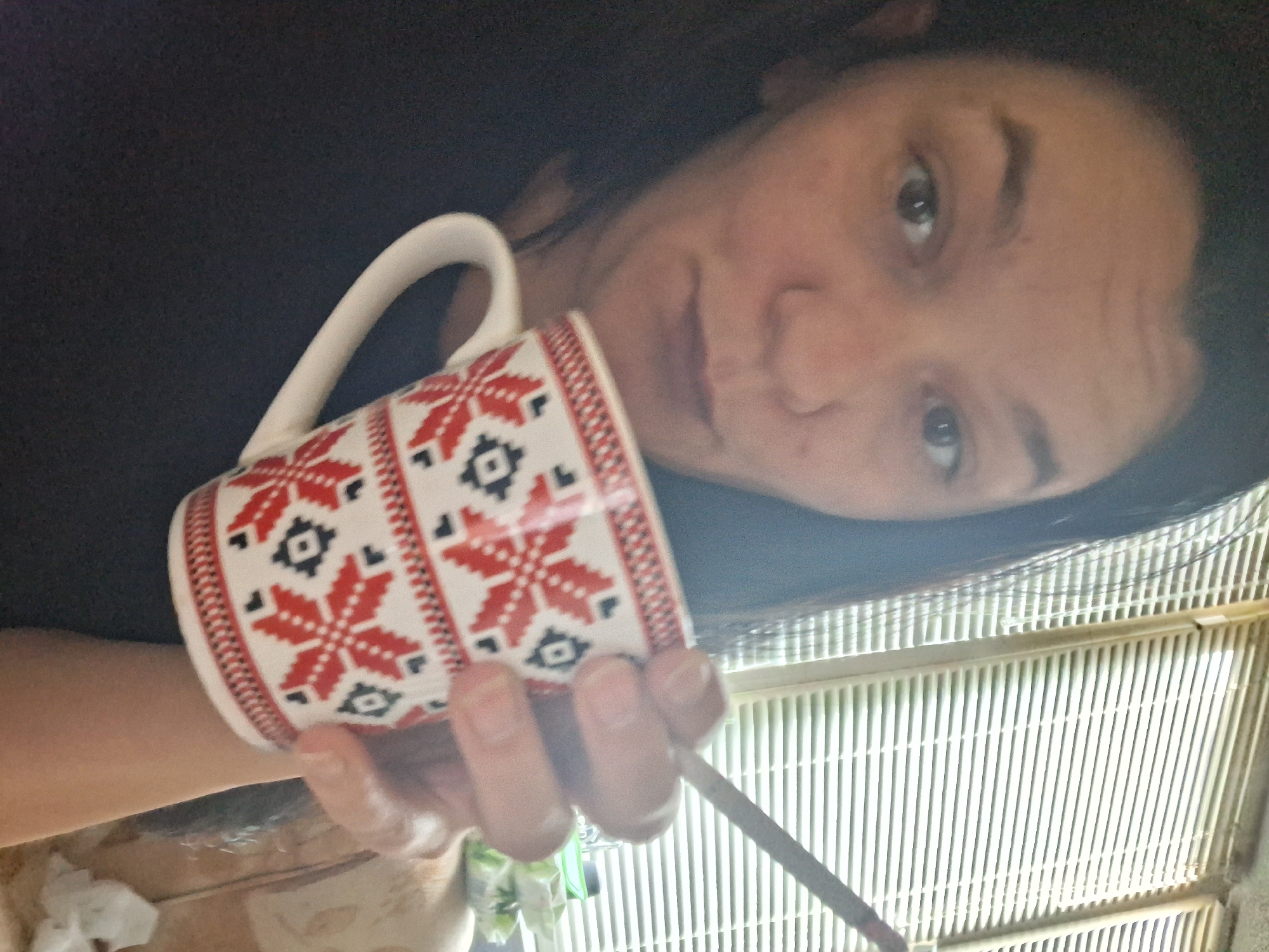 A woman with dark hair is holding a mug with a red and white snowflake pattern in her right hand. She is also holding a thin, cylindrical object, possibly a cigarette or a pen, between her index and middle fingers. The mug is positioned in front of her face, partially obscuring her mouth and chin. She is looking directly at the camera with her eyes open and appears to be indoors, near a window with venetian blinds. The background is slightly blurred, but suggests a domestic setting. There is no information to indicate this image is from Gotse Delchev, Bulgaria.