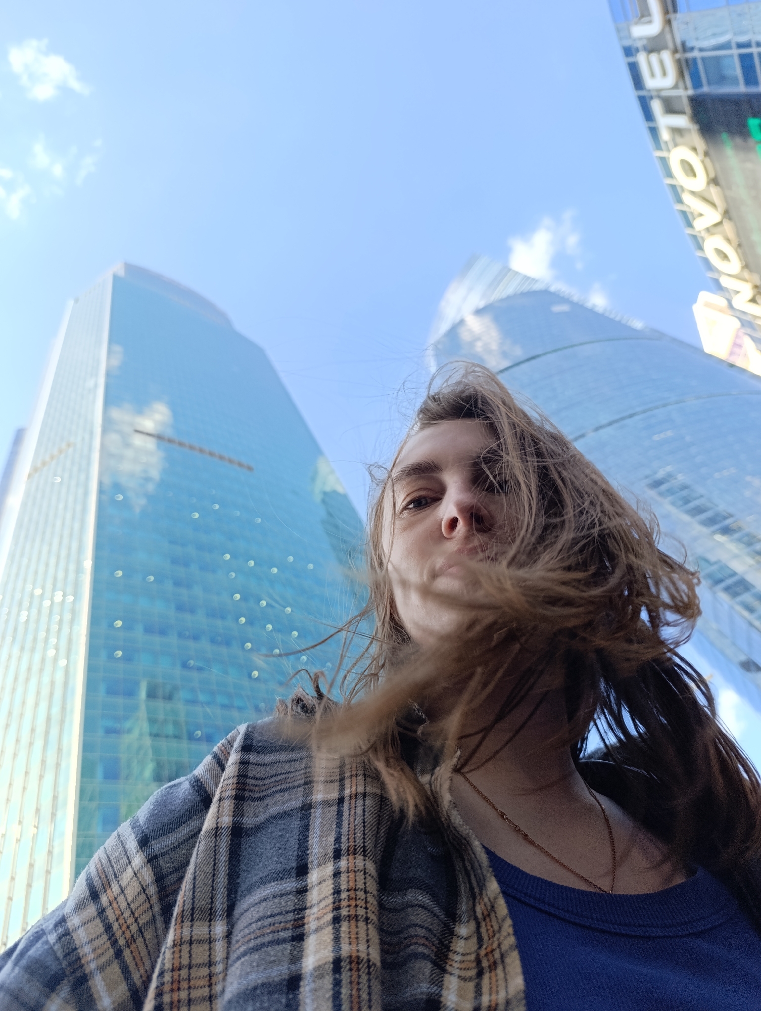 A low-angle shot captures a young woman looking down towards the camera, framed by towering glass skyscrapers against a bright blue sky. Her long, brown hair is swept to her right by the wind, partially obscuring her face. She wears a blue crew-neck top layered under a plaid shirt or jacket in shades of blue, beige, and brown, and a thin gold chain necklace is visible.
To the left, a tall, rectangular skyscraper with a blue reflective facade rises into the sky, mirroring scattered white clouds. To the right, another modern skyscraper, appearing more rounded at the top, also features a blue reflective surface. A white sign with the word "NOVOTEL" is prominently displayed on this rightmost building.
The scene takes place outdoors during the day under clear, sunny skies, suggesting a breezy day given the woman's hair. The extreme low-angle perspective emphasizes the scale and height of the urban architecture, creating a dynamic visual of a modern city setting.