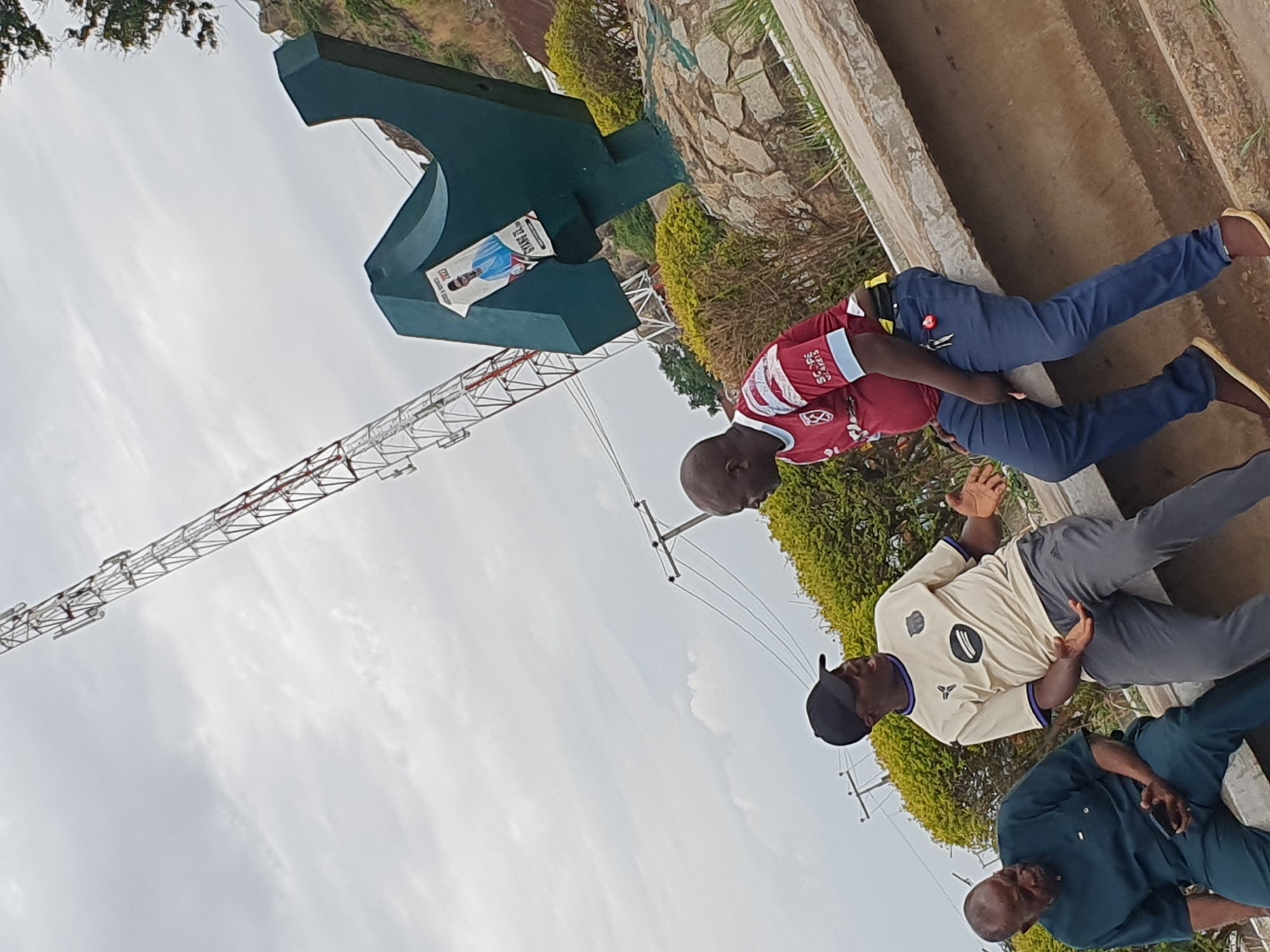 The image depicts three men near the Jos Plateau State Polytechnic sign in Kuru Dungus, Nigeria.
The man closest to the sign wears a maroon and white soccer jersey with blue pants and sandals. The next man is in a beige t-shirt, grey pants, and a black baseball cap. The third man wears a teal-colored dress shirt and darker pants.
Behind them is the large green Jos Plateau State Polytechnic sign with a poster attached. A communications tower is visible in the background. The environment includes greenery and a concrete structure. The sky is overcast.