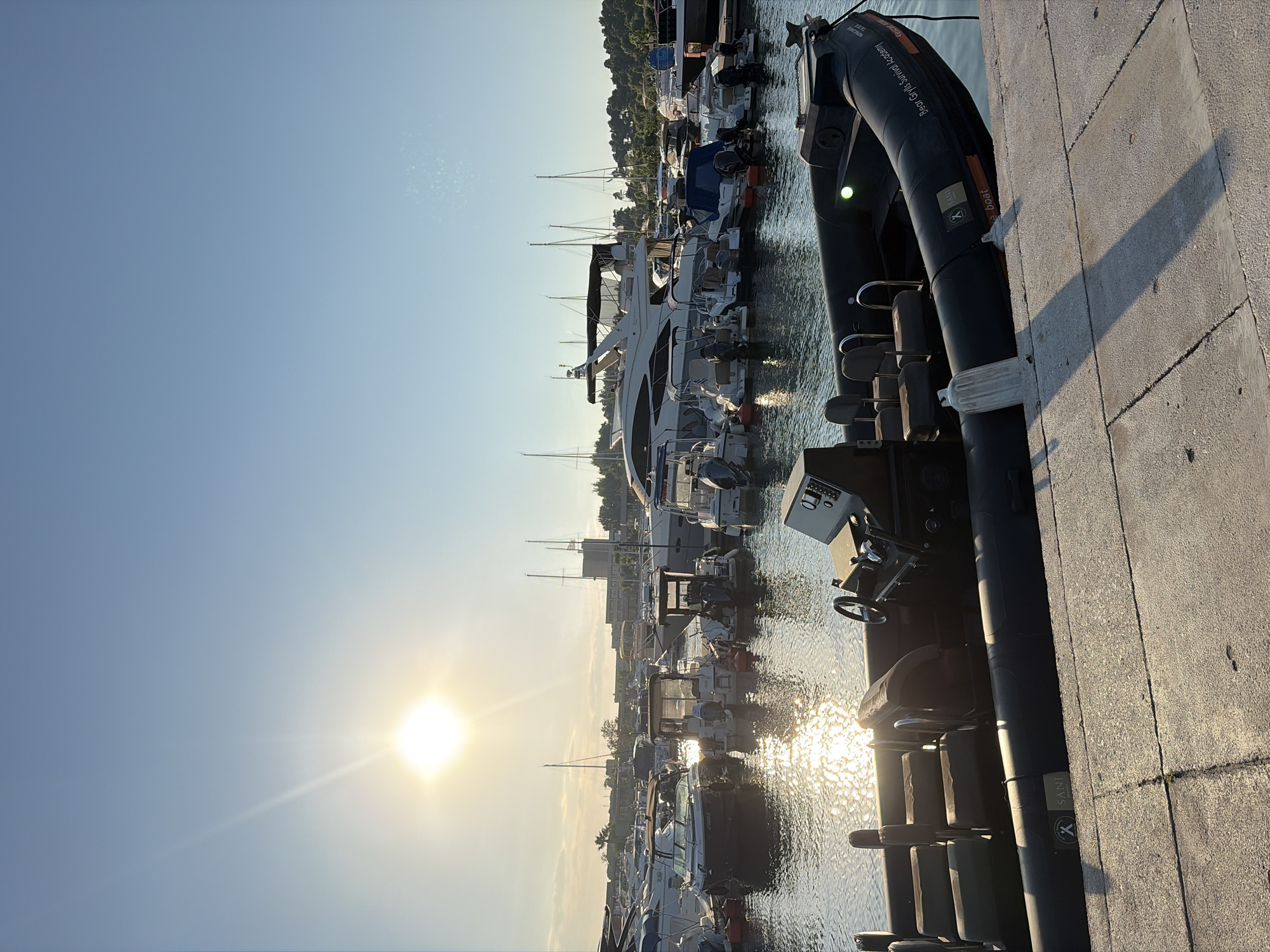 This image depicts a marina in the Kassandra Municipal Unit, Greece. The sun is low in the sky, casting a warm, golden light across the scene, suggesting it is either early morning or late afternoon. The weather appears clear, with a bright, cloudless sky.
The main subjects are a collection of boats docked in the water. These include various yachts and smaller vessels, some with protective covers. In the foreground, a large inflatable boat is partially visible, resting against a concrete walkway. This walkway is textured and shows a distinct shadow from the boat, indicating the direction of the sun.
The water is calm, reflecting the sunlight and the shapes of the boats. In the background, a tree-lined coastline with buildings can be seen, further establishing the location. The overall mood is peaceful and serene, characteristic of a quiet harbor.
Visible text on the inflatable boat reads: "Bear Grylls Survival Academy" and "SANI". The sun is a prominent element, shining brightly and creating a lens flare effect.