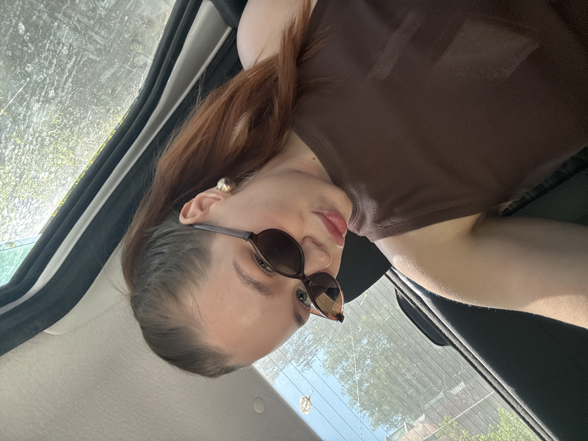 A woman is in the passenger seat of a car, wearing sunglasses and a brown top. The location is suggested as the city of Chaikovsky, Russia, but there is no definitive evidence of this in the image. The environment visible through the car windows includes trees and a building.