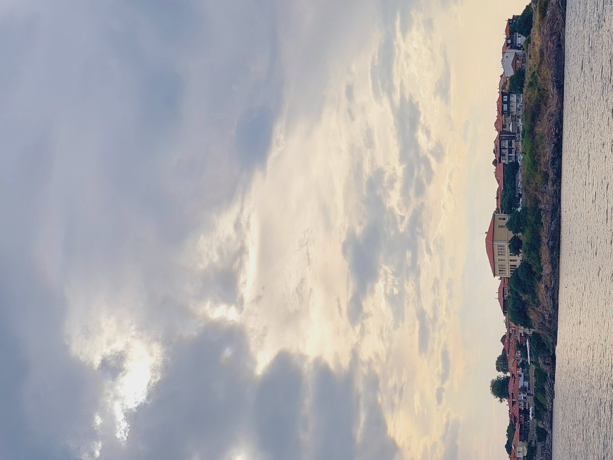 A wide, vertical, exterior shot captures the coastline of Sozopol, Bulgaria, under a dramatic, cloudy sky. The water in the foreground is calm and reflects the overcast sky with subtle ripples. On the rocky shore above, a cluster of traditional buildings with red-tiled roofs forms a picturesque townscape. The architecture is varied, with some houses featuring white or yellow facades and dark wooden accents, while others are more modern. Dense green foliage and trees are interspersed among the buildings, providing a lush contrast to the rocky terrain. The sky is filled with large, billowing clouds, with patches of brighter light suggesting the sun is attempting to break through. The overall impression is one of a serene coastal town on a somewhat moody day.