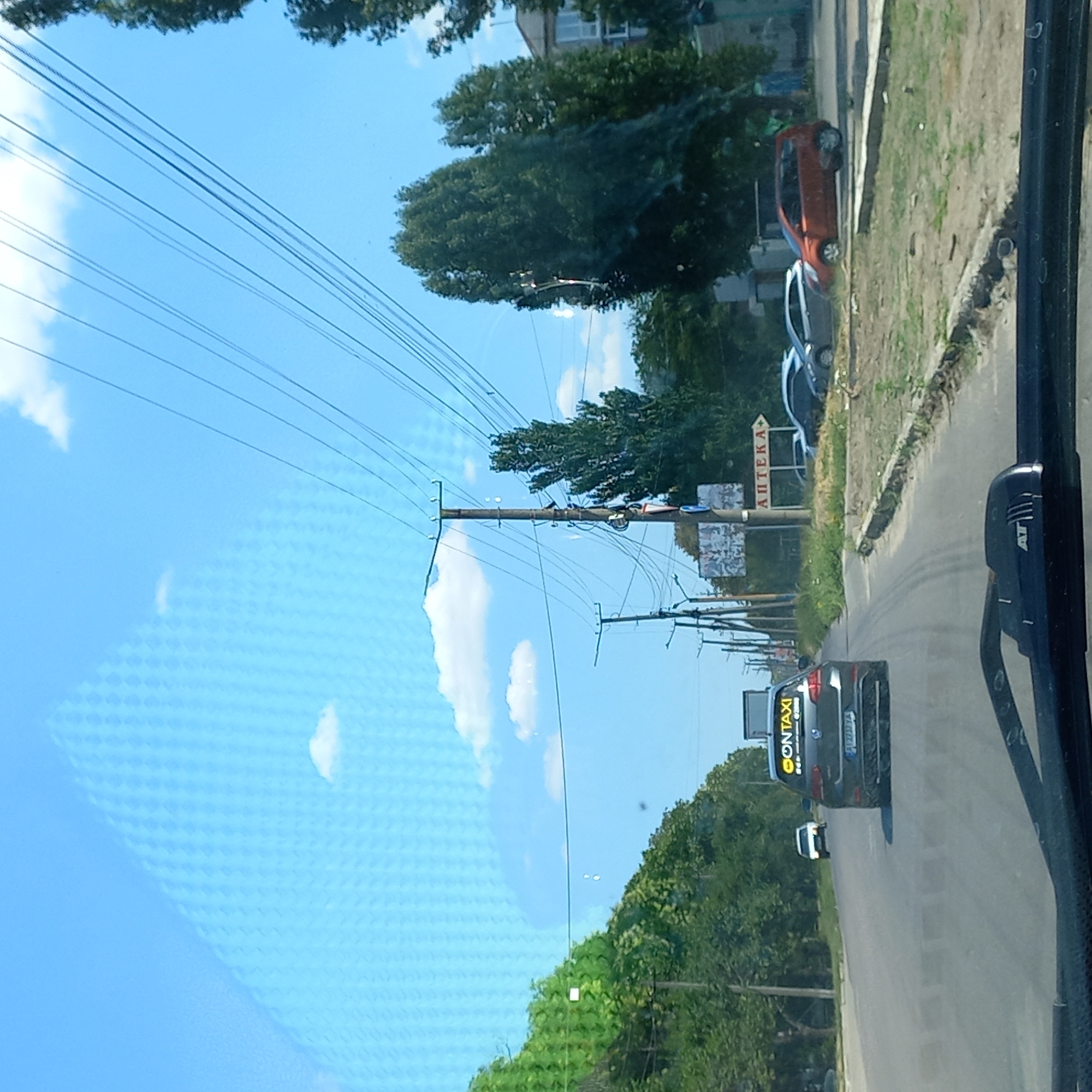 The image captures a street scene in Kropyvnytskyi, Ukraine, on a clear, sunny day. A gray Volkswagen Golf, identified as a "CONTAXI" vehicle by a yellow sticker on its rear window and a license plate reading "BI2733YA," is driving on a paved road. The car is in the right lane, moving away from the camera. In the distance, a white car is also visible on the road. A road sign with Cyrillic text, likely indicating "APTEKA" (pharmacy), points to the right. Overhead, numerous power lines crisscross the sky, supported by utility poles. The surrounding environment consists of trees lining the road, a grassy median strip, and some buildings in the background. The perspective suggests the photo was taken from inside another vehicle, looking forward.