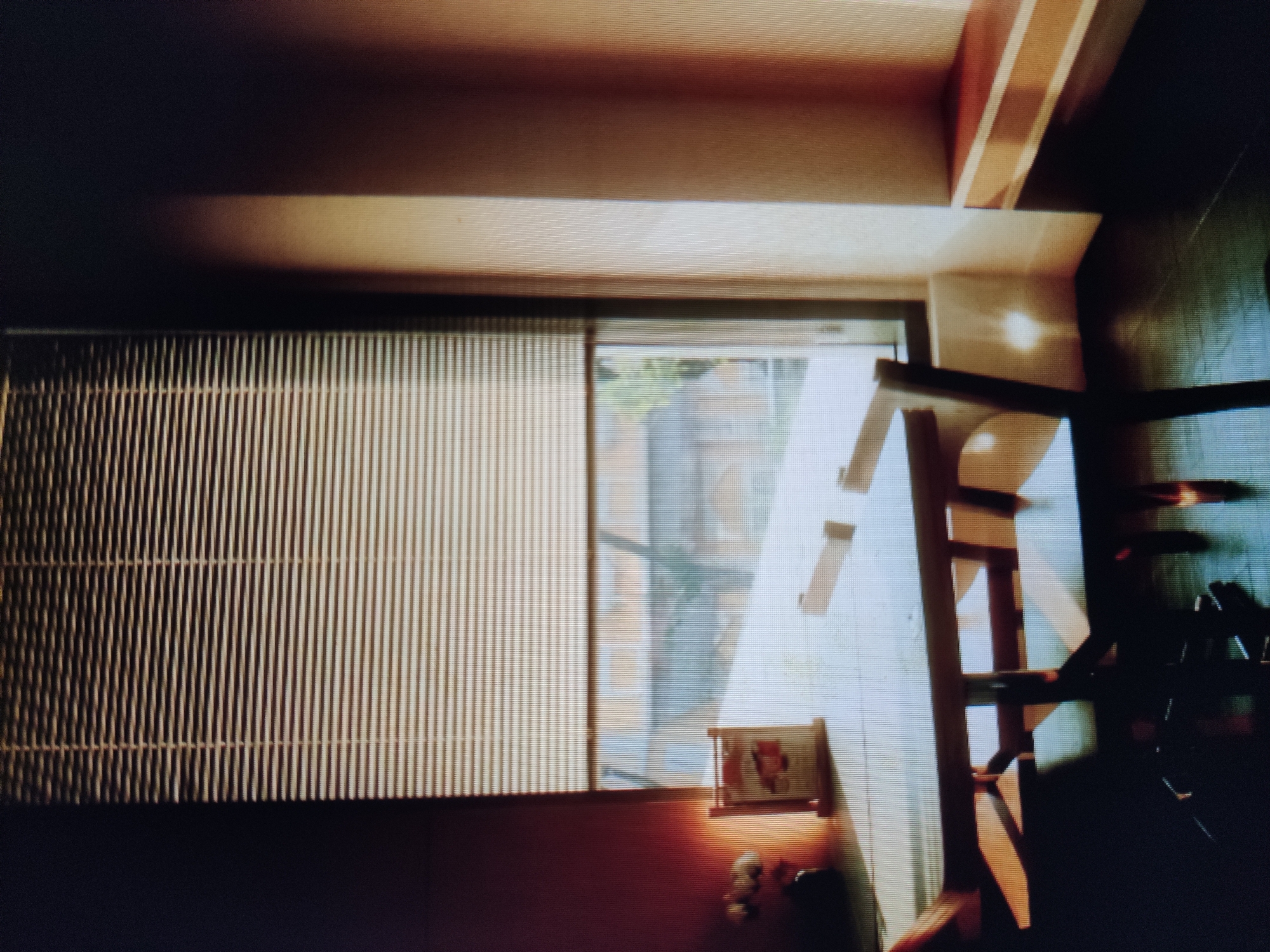 The image depicts an interior scene, likely a dining area or cafe, with a large window covered by horizontal blinds. Sunlight streams through the blinds, casting striped shadows on the wall and furniture. In the foreground, a wooden table with a light-colored top is set, with a small decorative item placed on it. Two chairs are partially visible, one in the foreground and another further back. The floor is dark, reflecting the light. The background outside the window appears to be a street scene with buildings and greenery, though it is blurred. The overall ambiance is one of quiet and warmth, with the sunlight creating a visually striking effect. The location is Unknown, Unknown.