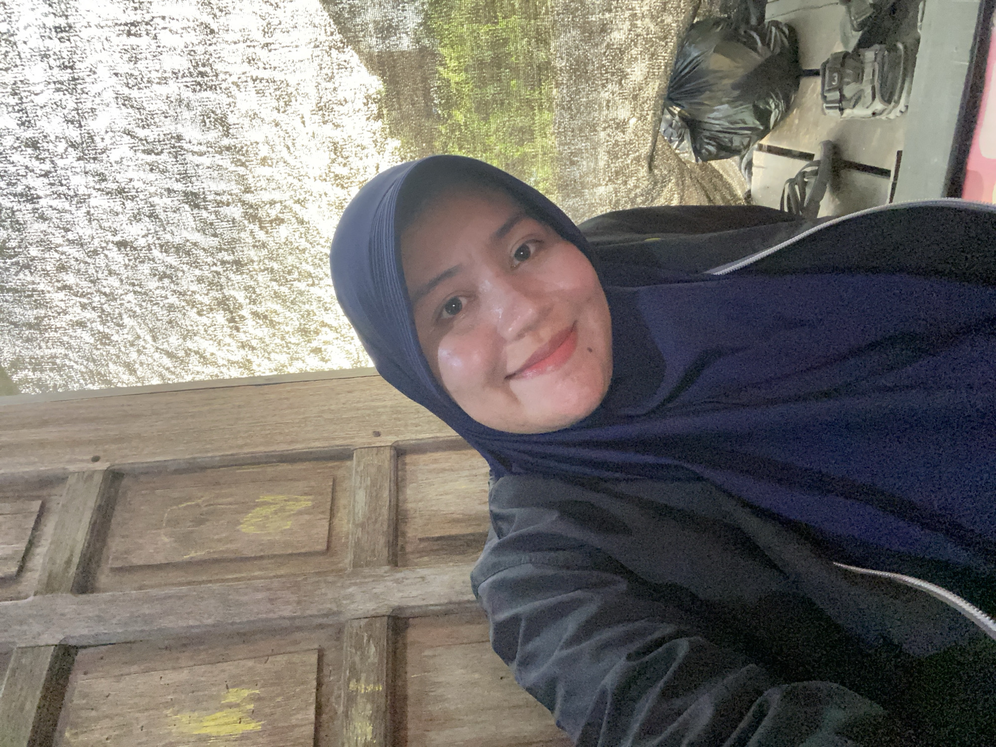 A woman wearing a dark blue hijab and a dark jacket poses for a selfie. Her face is illuminated, and she is smiling. She is positioned in front of a wooden door. To her right, a textured canvas screen is visible, and behind it, a black trash bag and a pair of shoes are on the ground. The overall setting appears to be outdoors.
