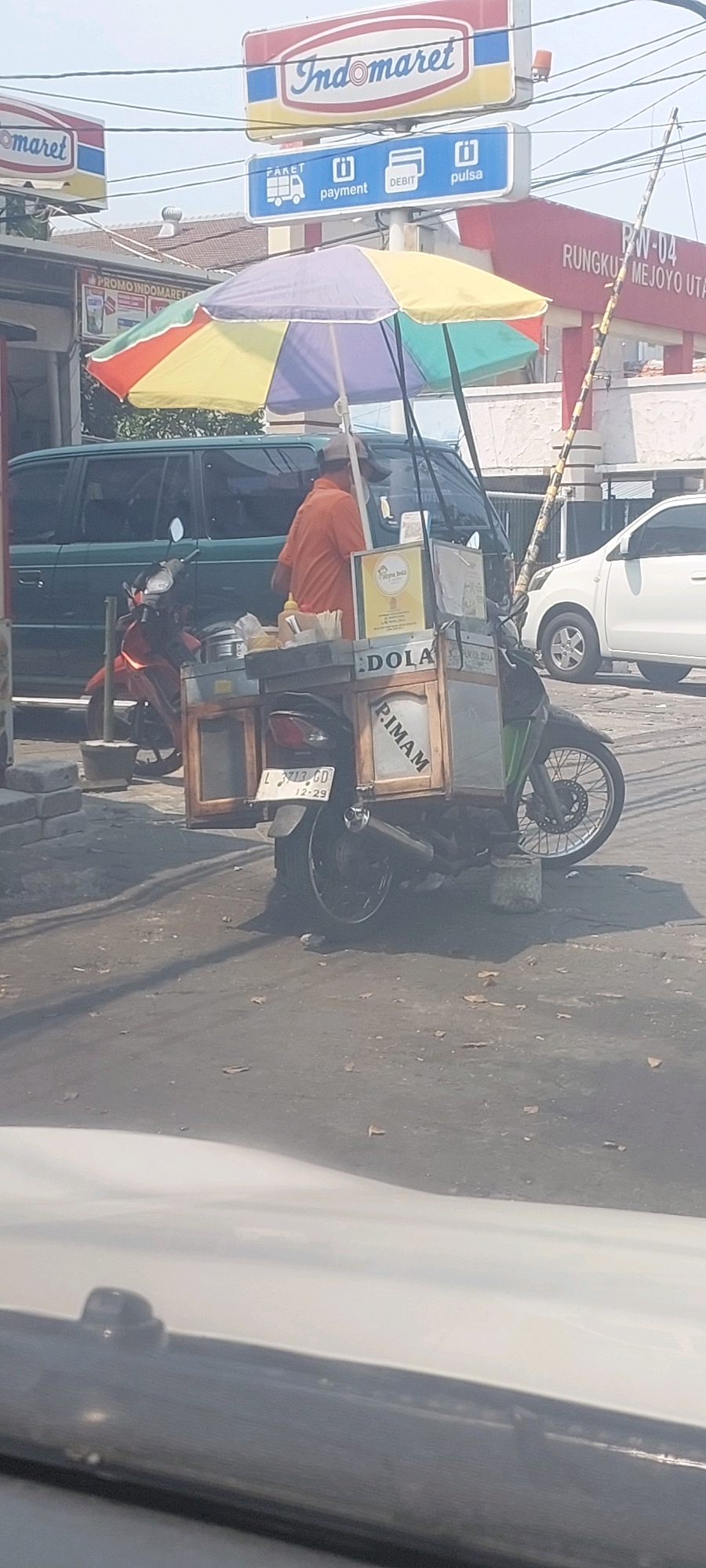In Surabaya, Indonesia, a vendor sells food from a cart attached to a motorcycle. The cart is colorful, with a multi-colored umbrella providing shade. The vendor, wearing an orange shirt, is partially visible. Behind the cart, a dark green minivan and a white car are parked on the street. In the background, the Indomaret convenience store sign is visible, indicating the location is likely near one of their branches. The scene appears to be a typical street-side vendor setup in a busy area.