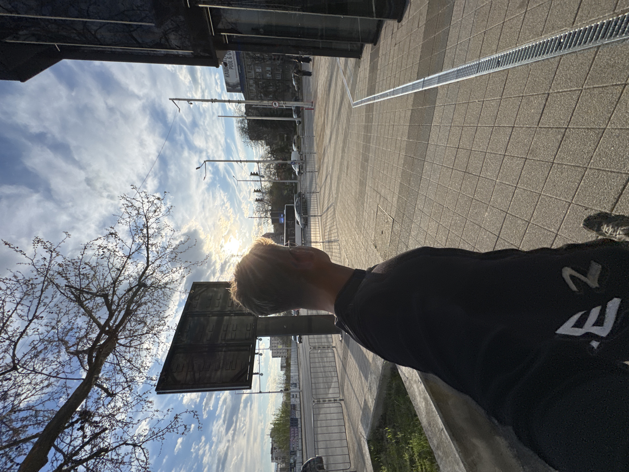 The photograph is taken outdoors in Plovdiv, Bulgaria. The perspective is from a low angle, looking upwards towards a bright sky with scattered clouds and the sun partially obscured by buildings.
In the foreground, a person with short blond hair, wearing a dark hooded sweatshirt with white lettering visible on the sleeve, is partially in the frame. The person appears to be looking out towards the scene.
The middle ground contains the urban landscape, including streets with parked and moving cars, buildings of varying heights and architectural styles, streetlights, and trees. A metallic drainage grate runs along the side of the street closest to the viewer. A large advertising billboard is positioned to the left of the person, adding to the urban clutter.
The background features the sky, which is filled with white and gray clouds. A bare tree with intricate branches stands in the left side of the frame, partially silhouetted against the bright sky.