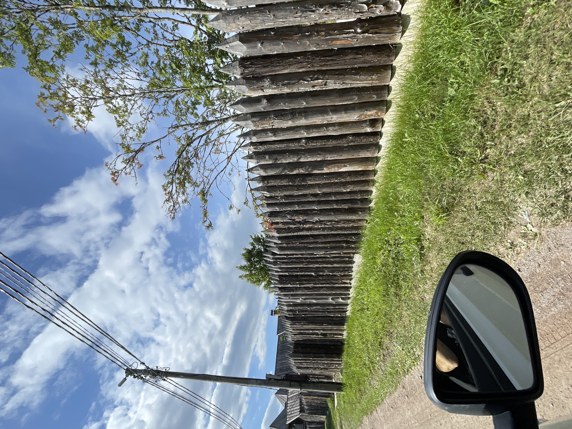 An eye-level, medium outdoor shot shows a dirt road next to a tall, sharpened wooden palisade fence on a sunny day with scattered clouds. The fence is made of rough-hewn logs, some showing signs of age and weathering. To the left of the fence, beyond a strip of green grass, are wooden buildings with peaked roofs, characteristic of older Russian architecture, suggesting a historical or rural setting within the city of Berozovo, Russia. A utility pole with multiple wires stands to the left of the fence, near the road. In the foreground, a car's side-view mirror reflects a person's arm and shoulder. The overall scene depicts a rustic roadside environment with elements of both nature and human construction.
