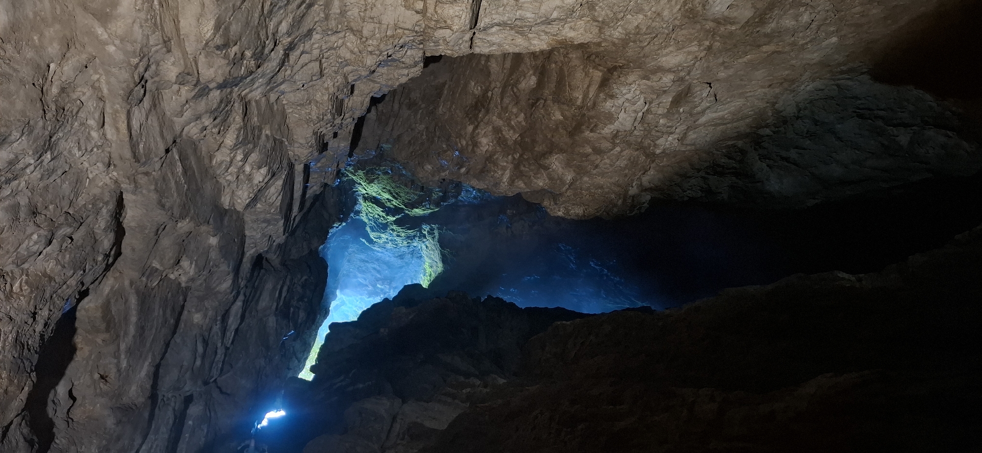 The image shows the interior of Trigrad Gorge, Bulgaria, a natural rock formation characterized by its rough, textured surfaces and deep crevices. Light streams in from an opening above, illuminating a section of the gorge with a vibrant blue glow. Some areas are shrouded in shadow, contrasting with the brightly lit sections. Moss or lichen clings to some of the rock faces, adding a touch of green to the predominantly grey and brown palette of the stone. The scene captures the dramatic and raw beauty of the natural environment.