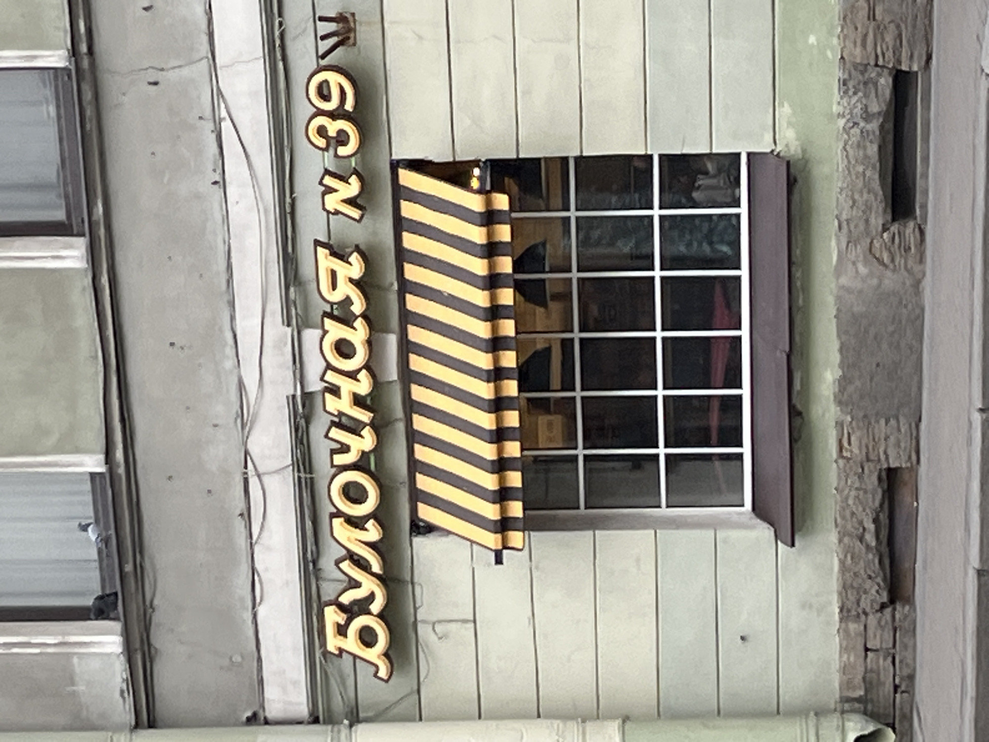 An exterior shot of a building in Saint Petersburg, Russia. The shot is angled upwards, focusing on a storefront facade. The sign reads "Булочная N 39," which translates to "Bakery No. 39." The sign is vertically oriented, with gold letters outlined in brown. The building facade is constructed of a pale green material, possibly painted wood panels. Next to the sign, there is a window with a brown frame, divided into 12 panes. A striped awning in yellow and brown is partially extended over the window. To the left of the sign is another window, higher up in the building, showing two pigeons. To the right of the window on the bottom, the side of the building has a rough, eroded appearance with exposed brick. A white pipe runs horizontally along the bottom of the image.