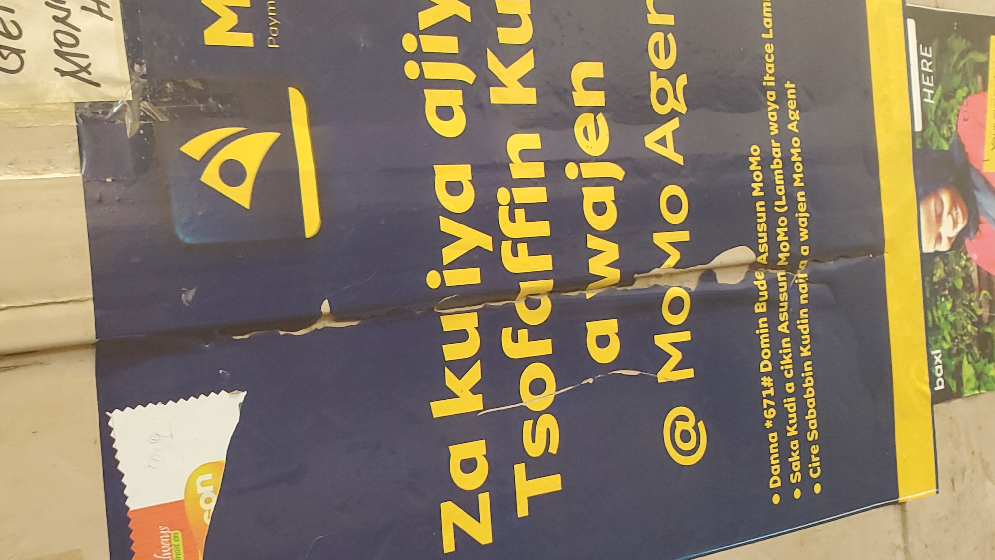 The image is a close-up, angled shot of a torn poster advertising MoMo Agent services. The poster is primarily dark blue with yellow text. The visible text includes "Za ku iya ajiy", "Tsofaifin Ku", "a wajen", and "@ MoMo Agen". There are also bullet points listing services: "Danna *671# Domin Bude Asusun MoMo", "Saka Kudi a cikin Asusun MoMo (Lambai waya itace Lamb", and "Cire Sababbin Kudina naita a wajen MoMo Agent". A yellow MoMo logo is visible on the upper right portion of the poster. In the bottom left, a small section of another poster or banner shows the word "baxi" and a smiling woman in the lower right quadrant of the frame, partially obscured. There is also a section of a light-colored surface at the top of the image, appearing to be a wall or another poster with handwritten text that is mostly illegible but includes "GET", "MONI" and "H". The poster is damaged with rips and tears, particularly along a vertical seam in the center. The lighting appears to be natural, suggesting it's an indoor or covered outdoor location. There is no indication of the specific location in Jalingo, Nigeria, within the image itself.