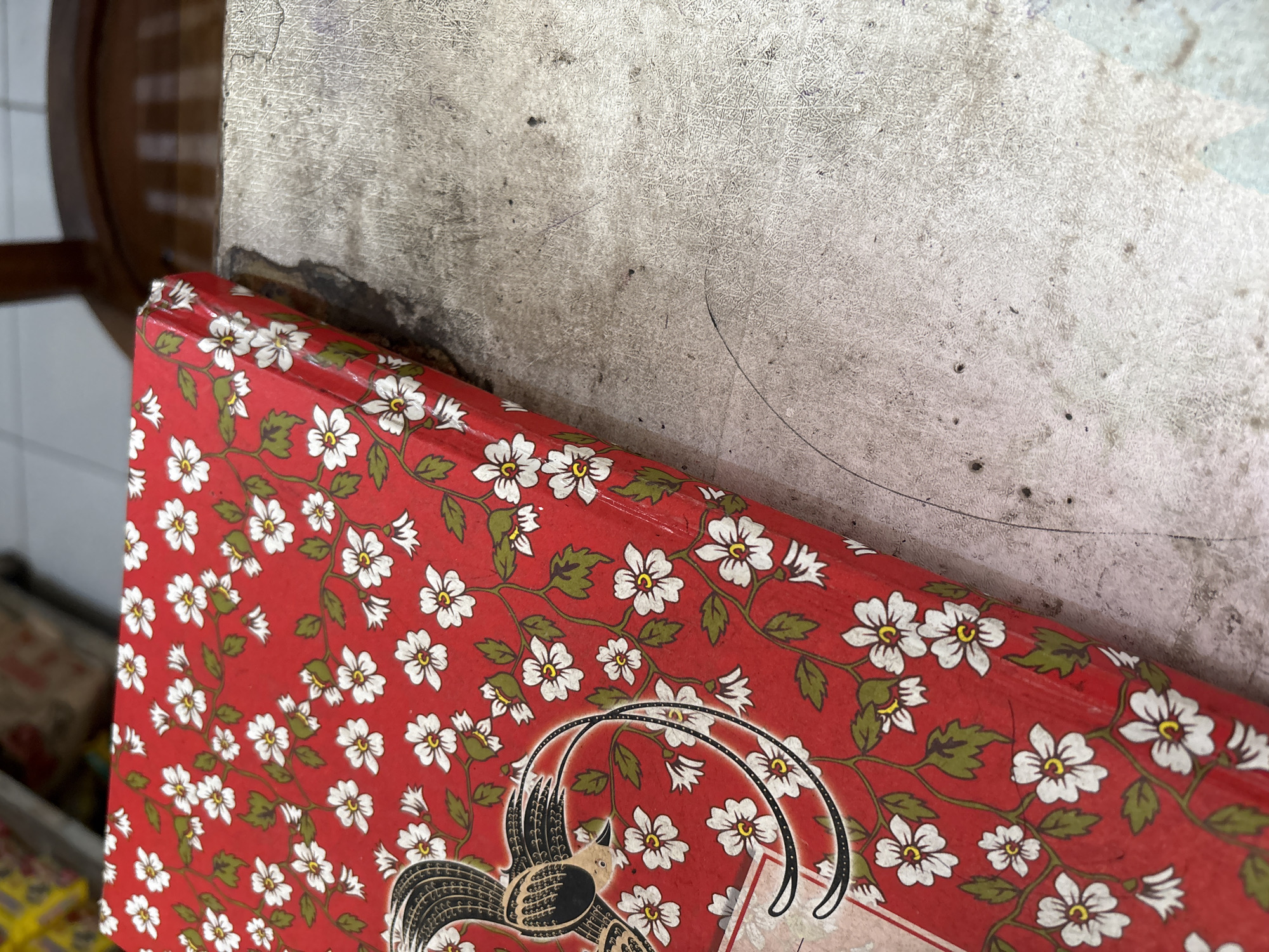 An overhead, close-up shot shows a red, floral-patterned box sitting on a textured, light-colored surface. The box, which appears to be made of cardboard or a similar material, features a repeating pattern of white flowers with yellow centers and green leaves, all arranged on a red background. A black and gold bird illustration is partially visible on the left side of the box. The textured surface beneath the box is worn and appears to be a tabletop or similar flat surface, with visible dust and smudges. The background is blurred, but suggests an indoor setting with hints of shelving and furniture. The image does not contain any identifiable location markers for Palimanan, Indonesia.
