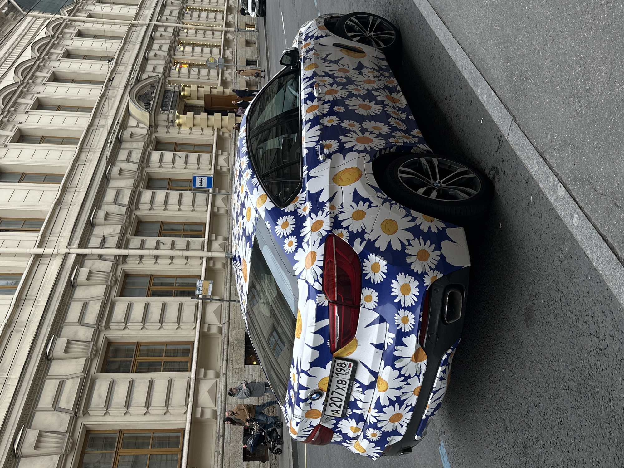 The image shows a parked BMW car, covered in a blue, white, and yellow floral pattern with daisies, on a street in Saint Petersburg, Russia. The car has a Russian license plate "A207XB 198 RUS". Beside the car, two pedestrians, a man in a brown jacket and another person with a stroller, are walking by. In the background, there's an ornate building with numerous windows and decorative architectural elements. A street sign is visible on the building, near the car. The setting suggests an urban area with classic European architecture.