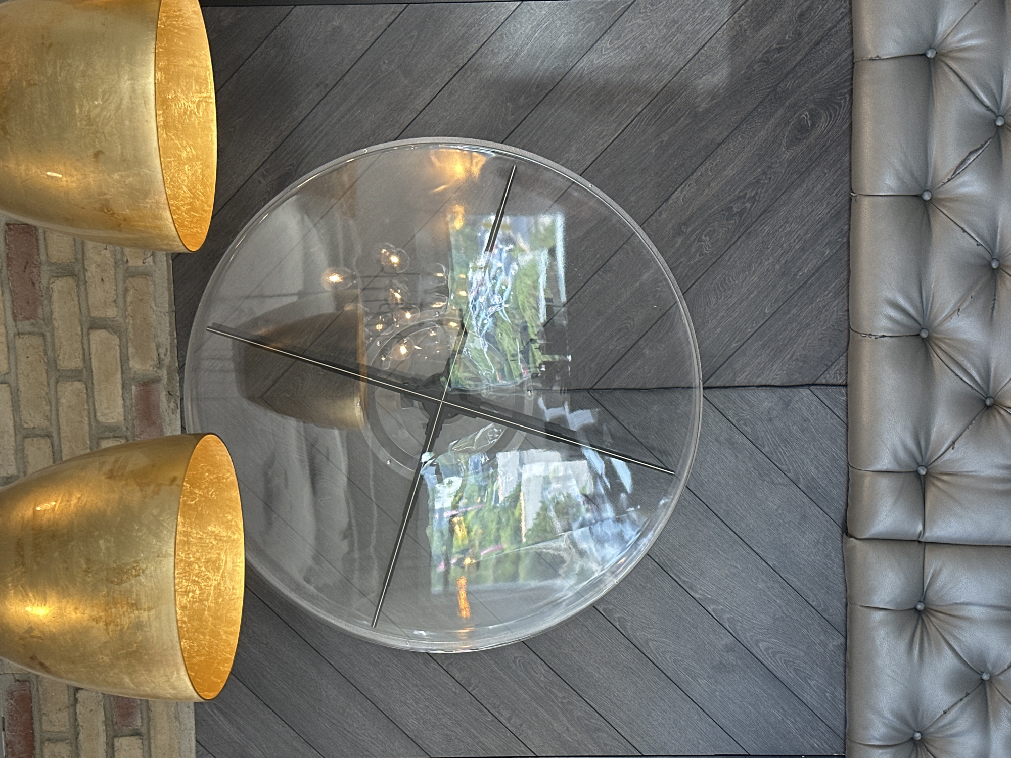A close-up, eye-level, vertical shot features a modern interior design with a large, clear, circular clock mounted on a dark, diagonally paneled wall. Two large, golden pendant lamps hang from above, their metallic sheen reflecting in the clock's surface. The clock's face is transparent, revealing a reflection of what appears to be an outdoor scene with greenery and a body of water, as well as a chandelier with multiple bulbs. The wall paneling has a wood-grain texture. Below the wall, a segment of a tufted, grey leather sofa is visible, showing the button-tufted pattern. The image does not provide enough information to determine the specific location in Coquitlam, Canada.
