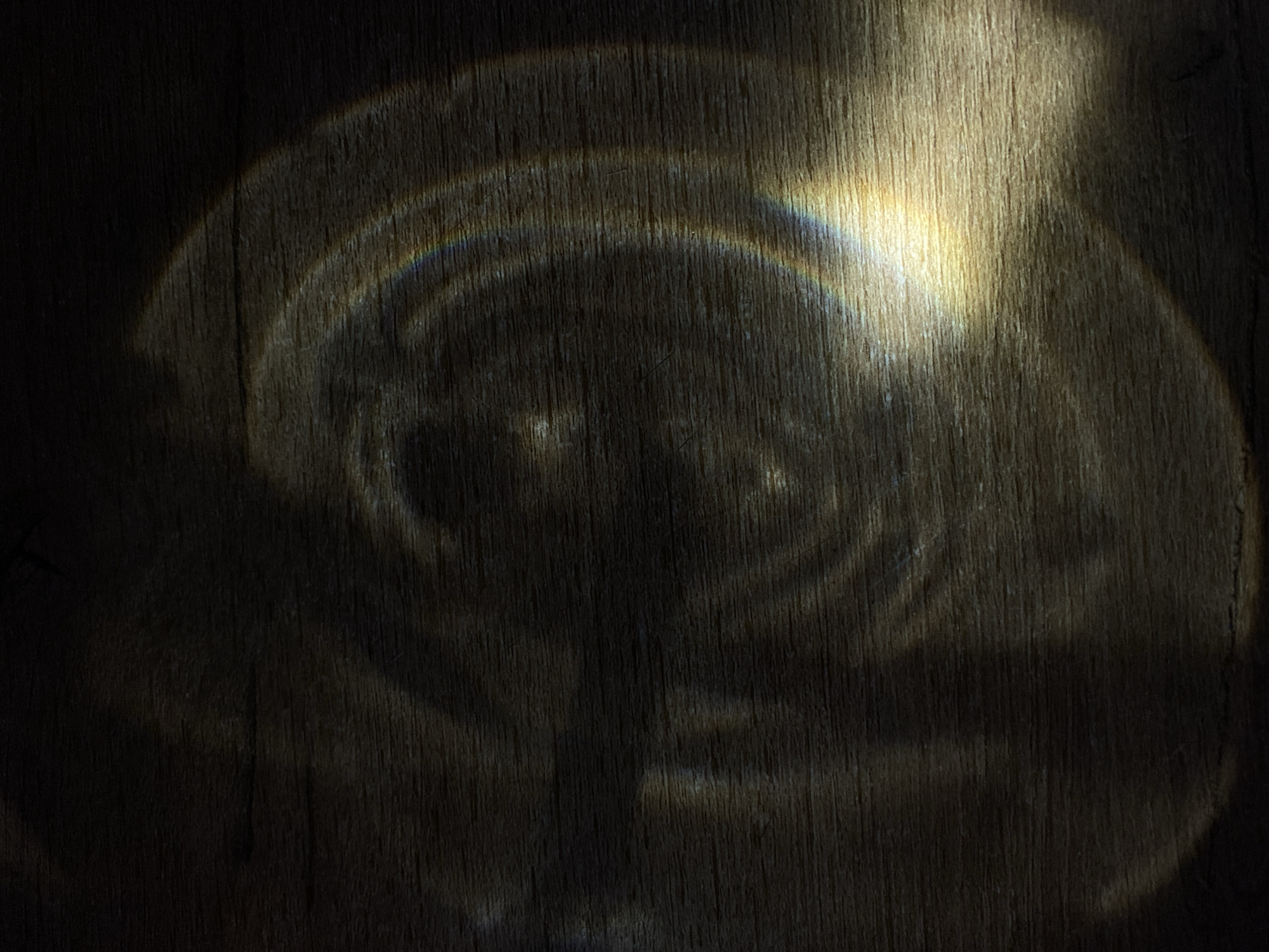The image displays a dark, textured wooden surface featuring horizontal grain patterns and minor scuffs. A distinct light caustic pattern is cast upon this surface, characterized by a series of concentric and irregular arcs of light. The brightest segment of this pattern, located towards the lower right, exhibits a clear prismatic effect, refracting light into a spectrum of colors including blue, green, yellow, and red. The surrounding wooden surface is largely in shadow, emphasizing the luminous projection. No individuals, objects beyond the surface itself, or active events are visible. This scene was captured in Chaikovsky, Russia.