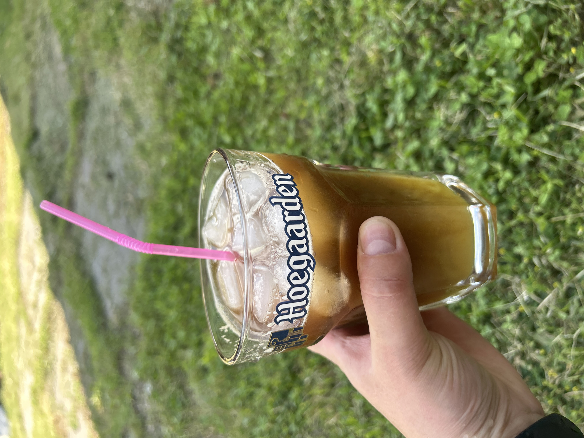 A hand is holding a glass of iced coffee with a pink straw. The glass has the Hoegaarden logo on it. The background is a grassy area. There is no information about the location in Bila Tserkva, Ukraine.