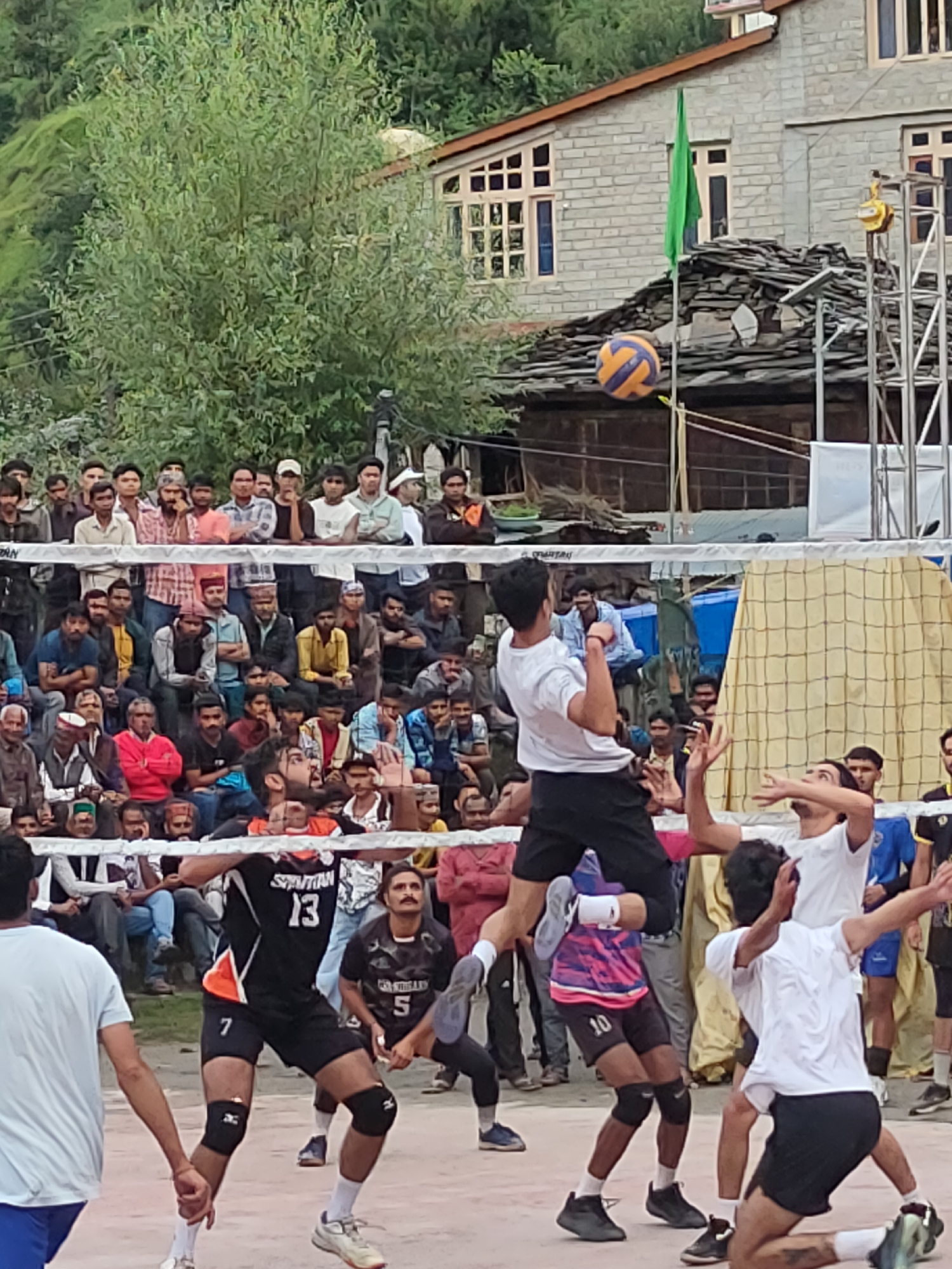 This image captures a volleyball match in progress in the mountainous region of Burwa, India. The scene is set outdoors, with a volleyball net dividing the court. Several players are actively engaged in the game. In the foreground, one player in a black jersey with orange accents, numbered 13 and 7, is mid-air, reaching for the ball. Another player in a white t-shirt is positioned to receive the ball. A third player, wearing a pink and blue patterned jersey with the number 10, is also actively involved in the play. Behind the players, a crowd of spectators is gathered, some seated and others standing, observing the match. The background reveals a rustic environment, with traditional stone buildings and steep, tree-covered hills characteristic of the Himalayan landscape. A green flag is visible on a pole near one of the buildings. The overall atmosphere appears to be one of community engagement and sporting enthusiasm in a rural Indian setting.
