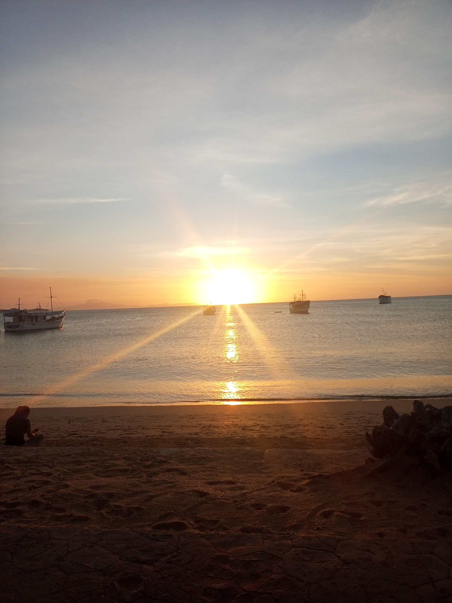 The image captures a sunset over a body of water, likely a bay, with a sandy beach in the foreground, located in Juan Griego, Venezuela. The sun is low on the horizon, emitting a bright, golden light that creates a prominent sunburst effect and a reflective path across the water directly towards the viewer. The sky exhibits a gradient from pale blue at the top, transitioning to orange and gold near the horizon, with scattered, thin clouds.
Several boats are visible on the water. A larger, light-colored boat with two masts and rigging is anchored on the left side of the frame. Two additional, smaller vessels are visible further out, with one positioned within the sun's direct reflection and another to its right.
The immediate foreground is a dark, sandy beach, darkened by the backlighting. Footprints are discernible on the sand, and some cracked ground patterns are evident in the lower right. A single individual is seated on the sand in the lower-left quadrant, presented in silhouette, facing the water and the setting sun. This person appears to be observing or interacting with a small object held in their hands. To the far right, a dark, irregular cluster of objects, resembling driftwood or rocks, rests on the sand.